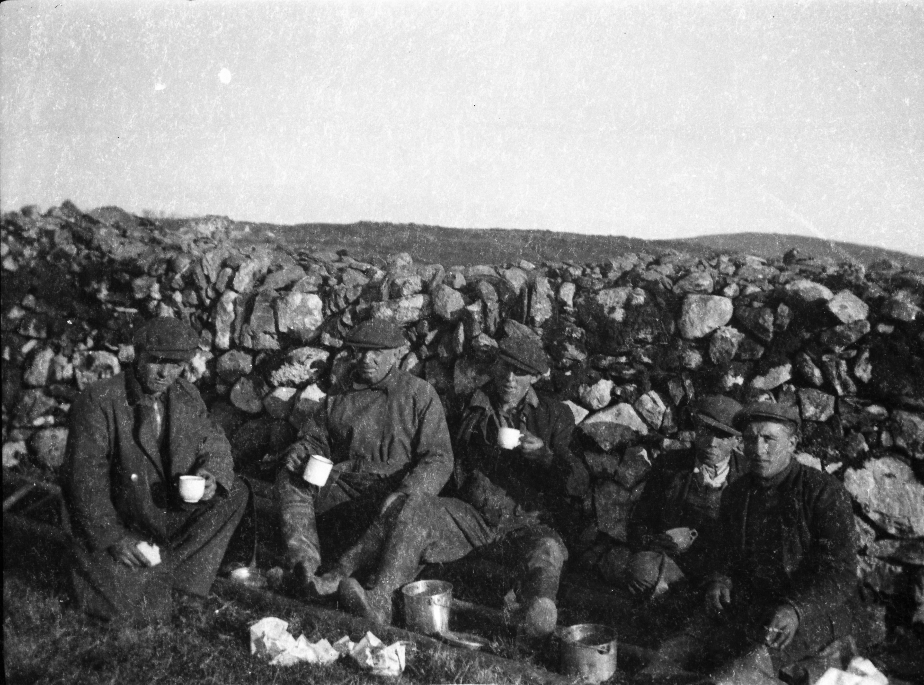 Black-and-white photograph showing five electricity network construction workers resting beside a stone wall and sharing refreshments in the countryside.