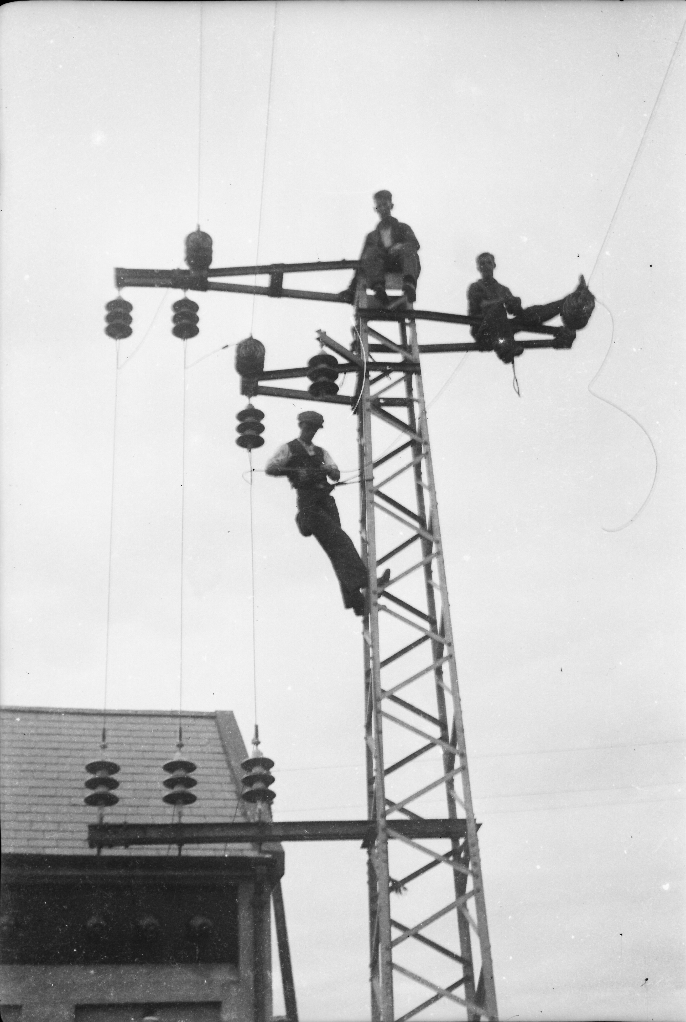 Black-and-white photograph showing workers carrying out construction work on a steel electricity tower beside a substation building.