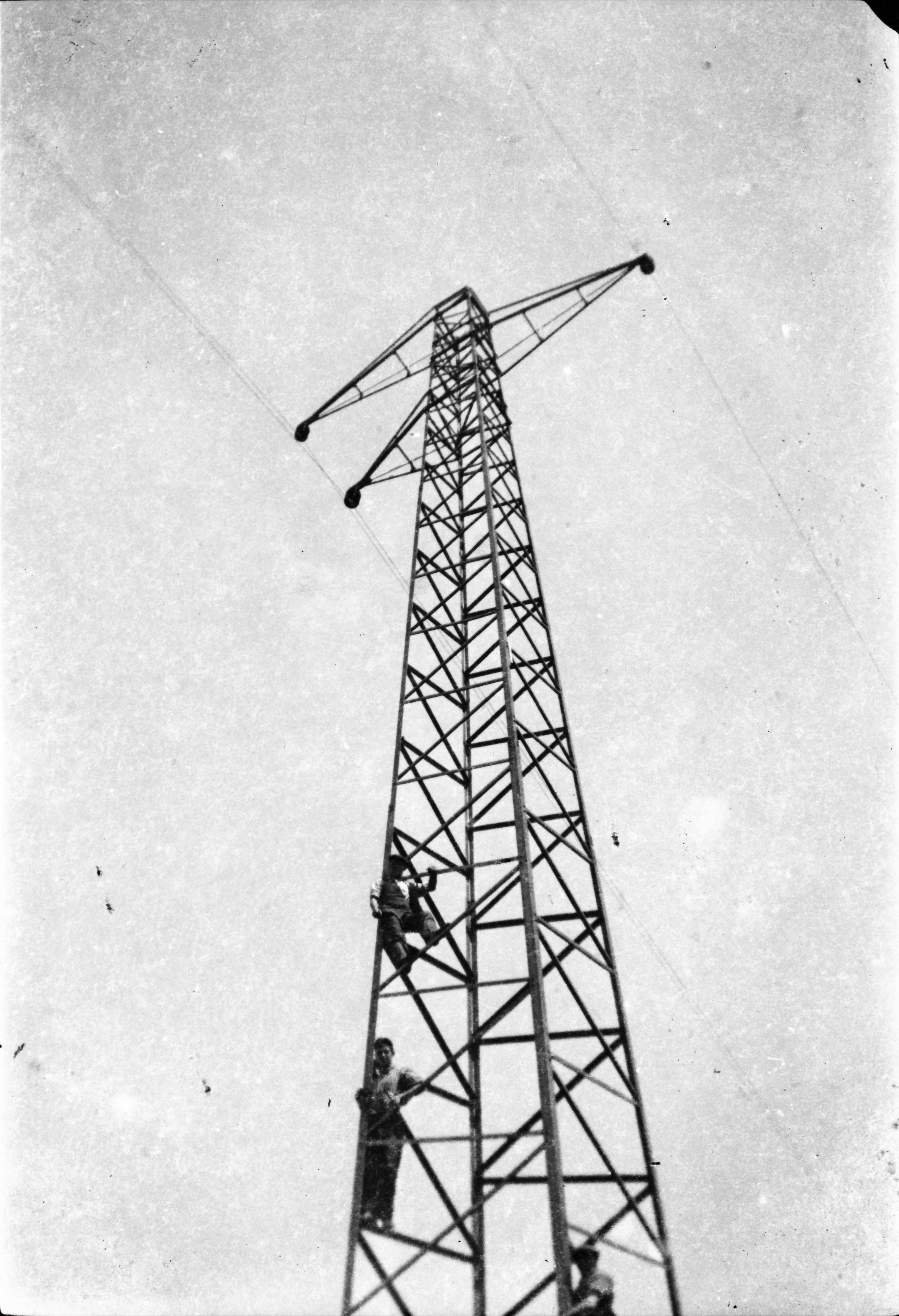 Black-and-white photograph showing workers ascending a steel transmission tower during electricity network construction, viewed from ground level looking upward.
