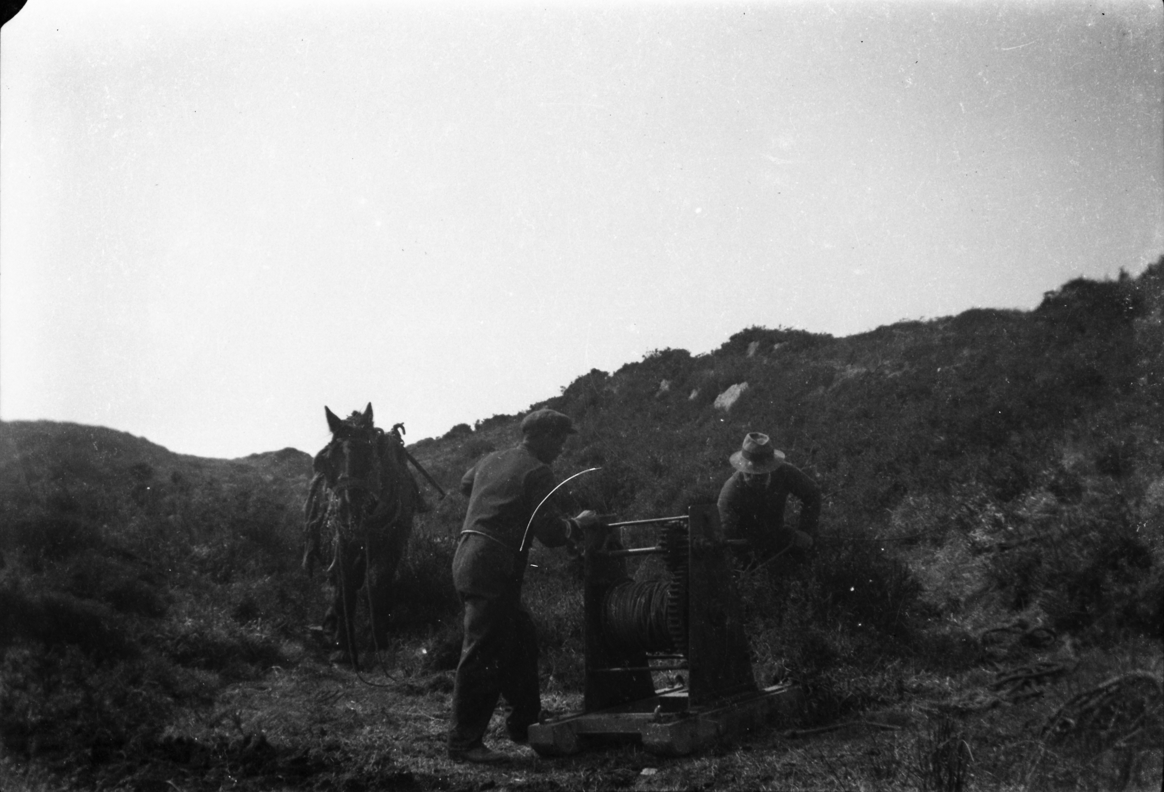 Black and white photograph showing two workers handling a cable reel mounted on a frame in open countryside, while a harnessed horse stands beside the equipment among heather-covered hills.