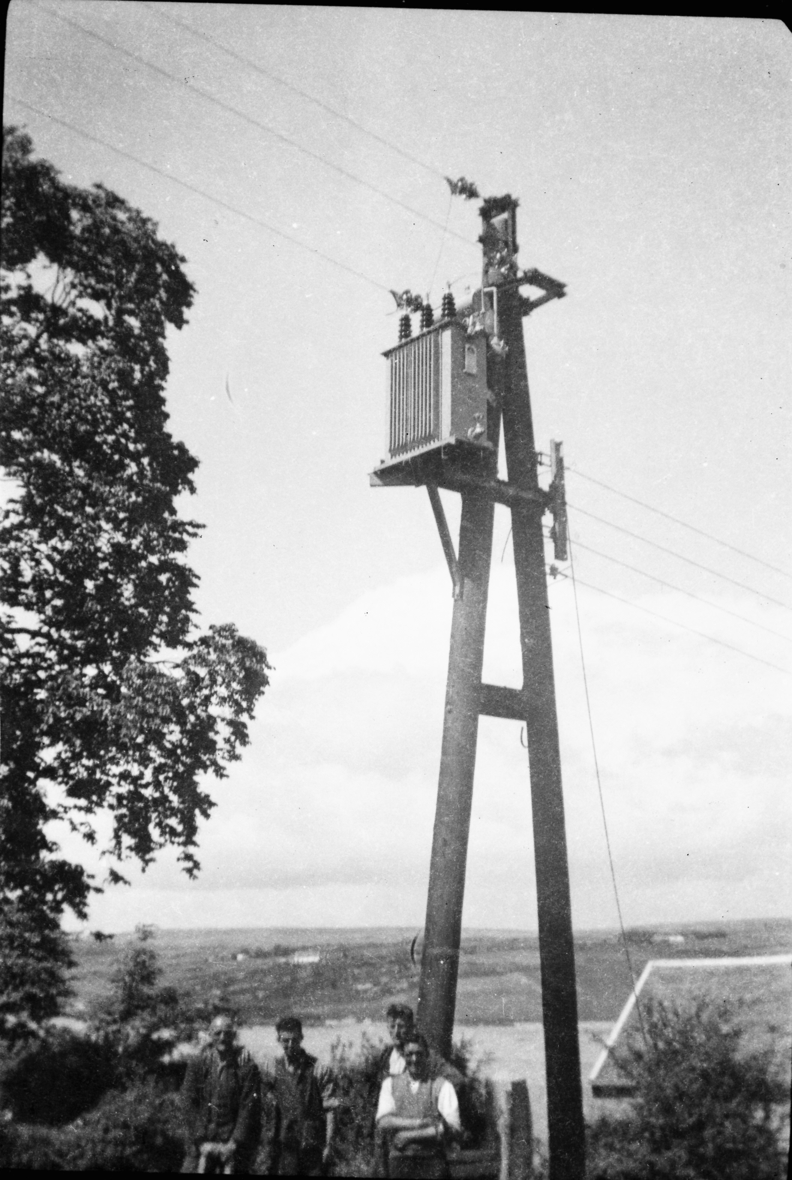 Black-and-white photograph showing an electricity transformer mounted on a wooden pole structure, with four people gathered beneath it.