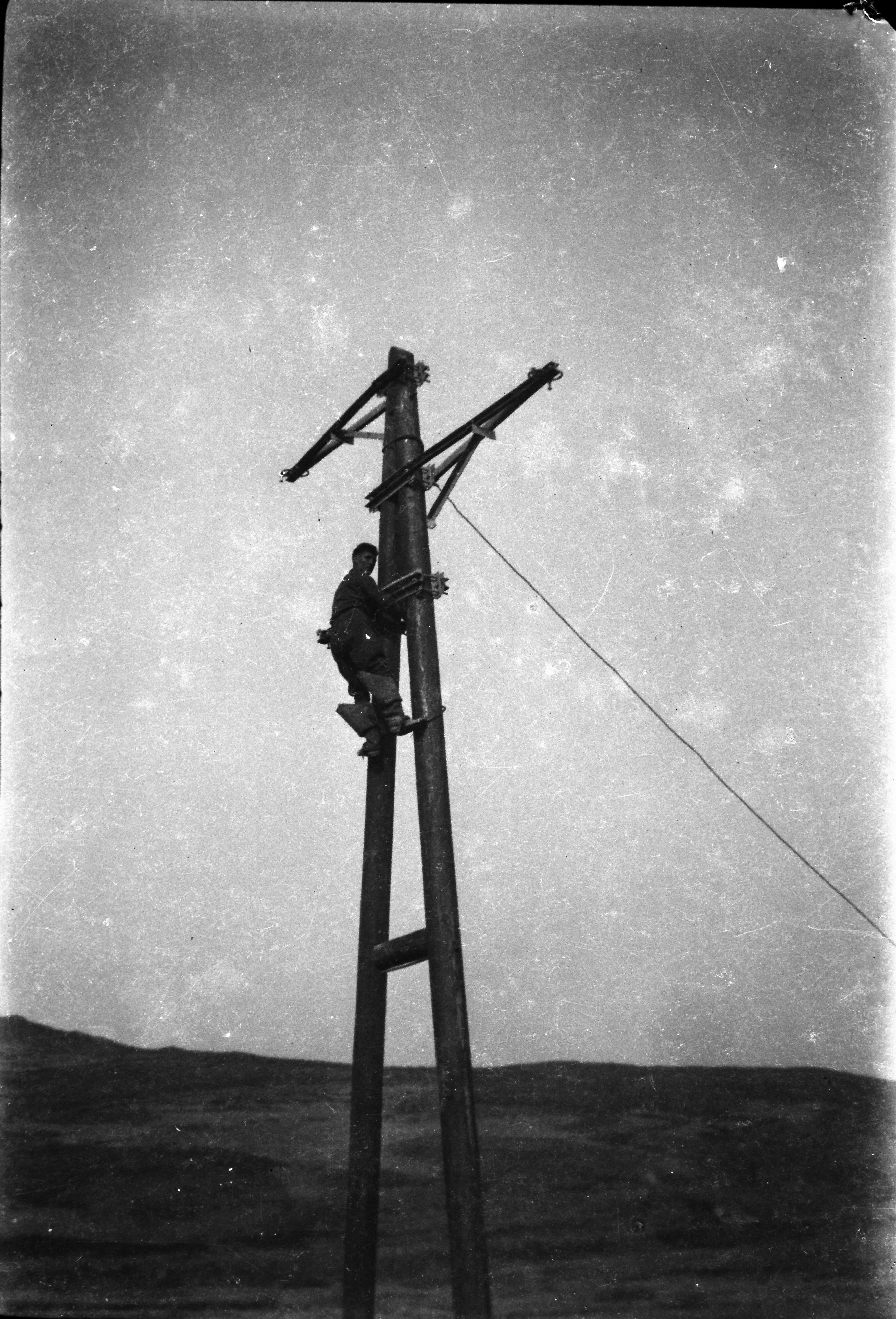 Black-and-white photograph showing a line worker ascending a wooden electricity support structure during construction of the rural electricity network.