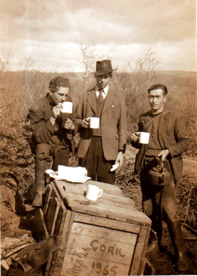 Three ESB workers taking a tea break outdoors beside a wooden crate, each holding a mug, with a kettle and rural landscape visible around them.