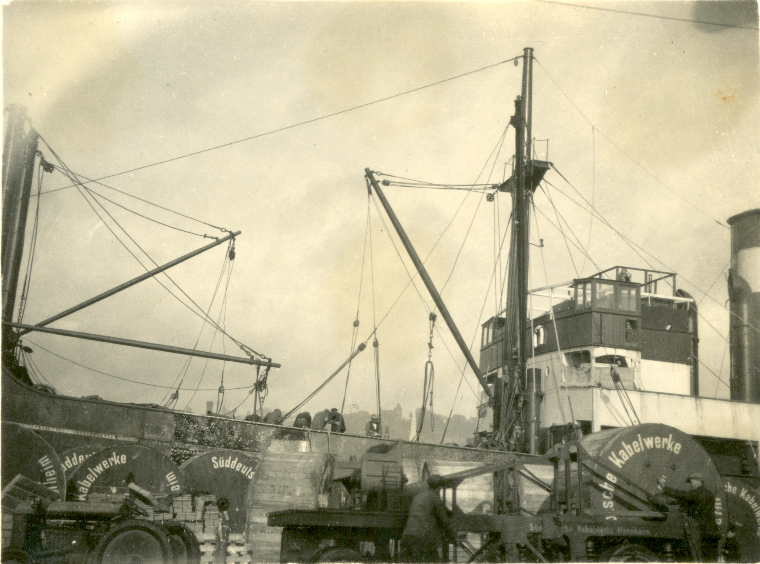 Black-and-white photograph showing large cable drums being unloaded from a cargo ship at a dock. Cranes and lifting equipment are used to move the drums, which contain electricity distribution cables for power network construction.