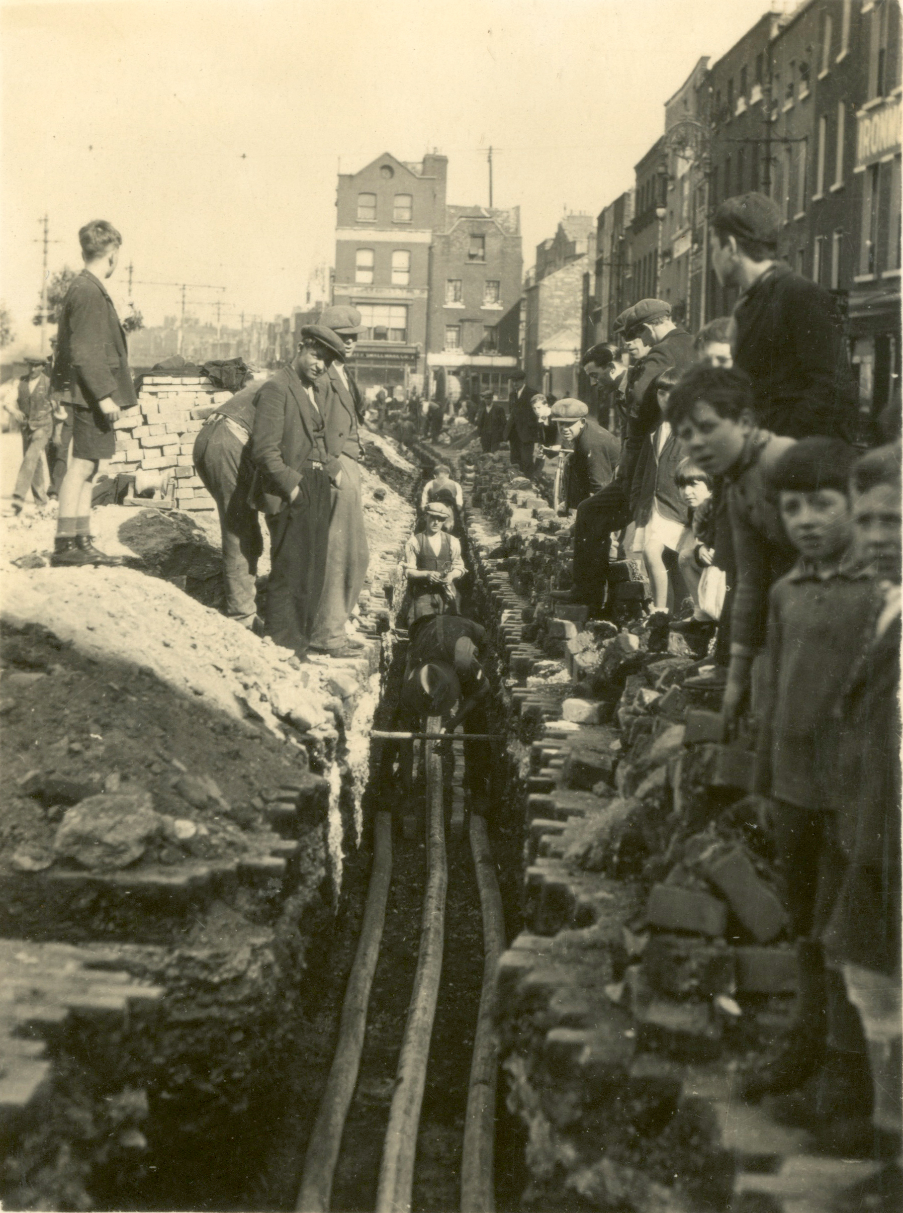 Black-and-white photograph showing workers installing underground electricity distribution cables in a street trench while local residents watch from either side of the excavation.