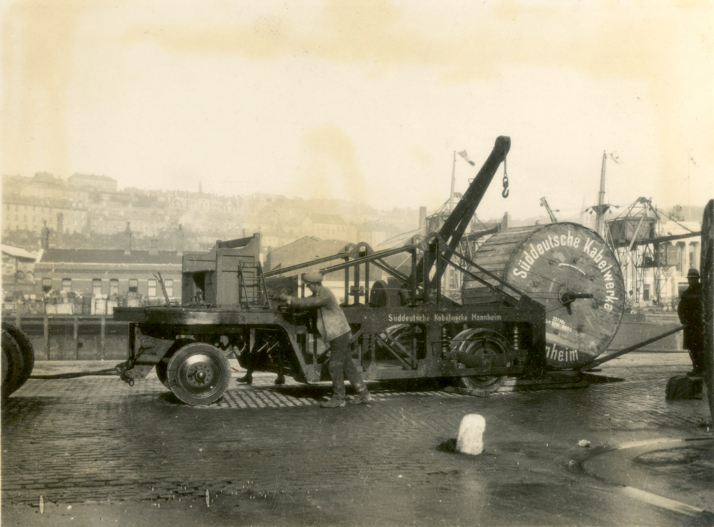 Black-and-white photograph of a large cable reel mounted on a trailer being positioned by workers beside a waterfront. Industrial cranes, buildings, and harbour structures are visible in the background.