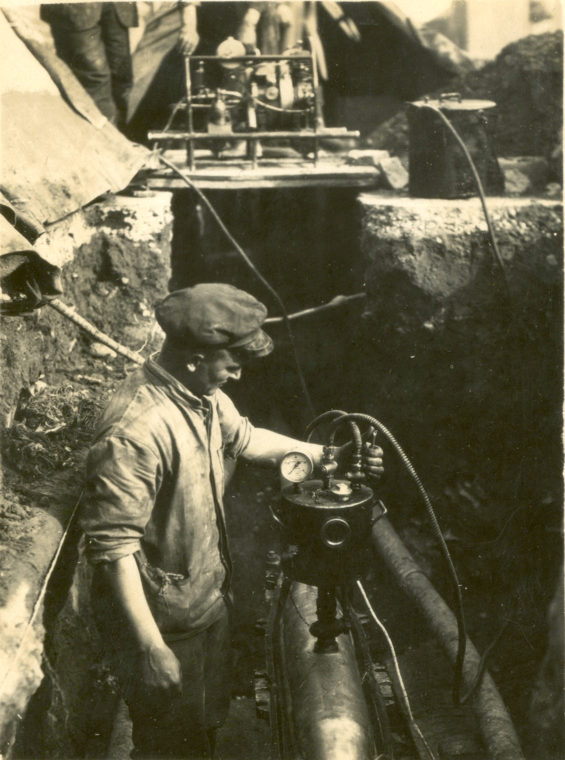 Historic black-and-white photograph showing a worker in a utility trench testing or monitoring an underground electricity cable using specialised equipment during the installation of the electricity distribution network.