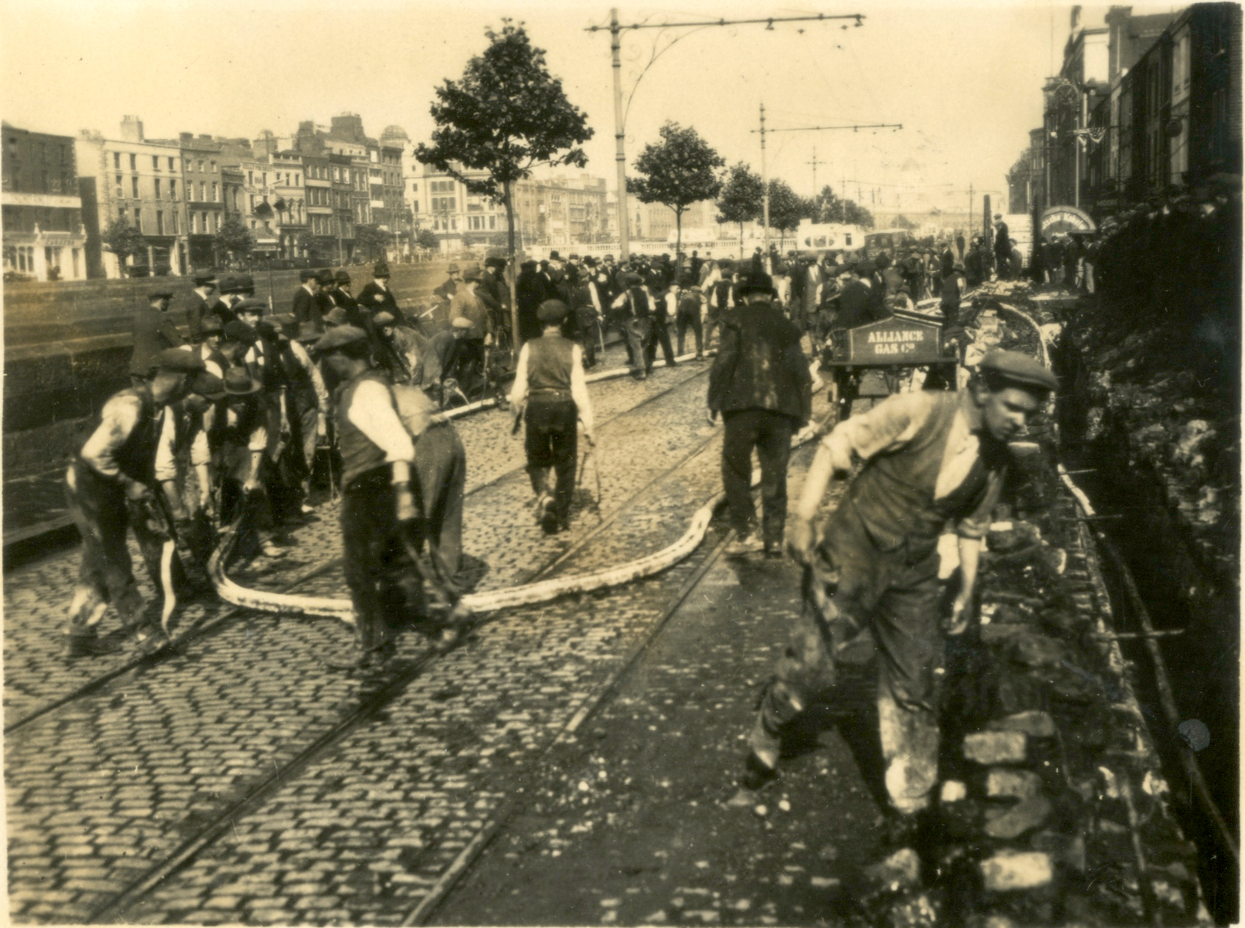 Black-and-white photograph showing workers laying electricity distribution cables along a cobbled city street. A large workforce is gathered around the cable installation site, where cables are being positioned beside tram tracks.