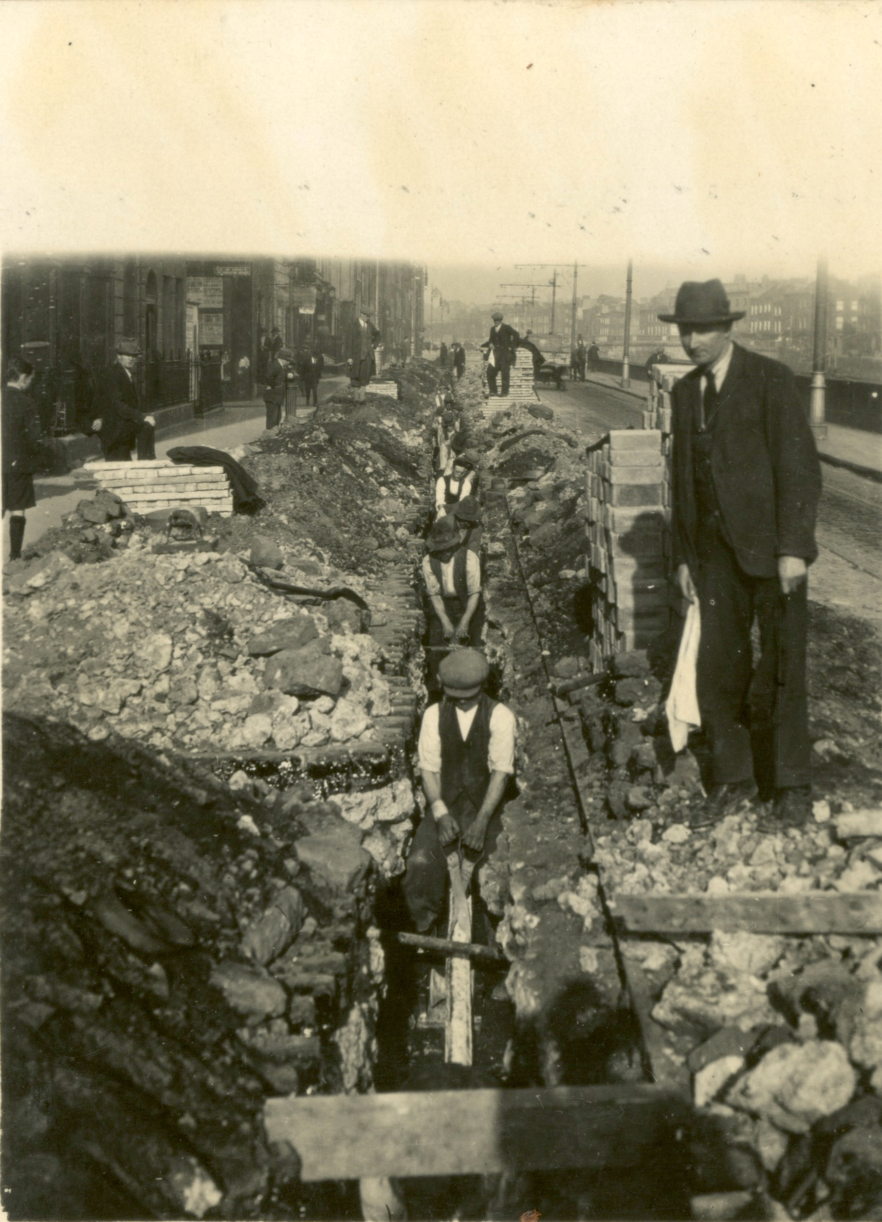 Black-and-white photograph showing workers laying an electricity distribution cable in a trench excavated along a city street. Construction materials line the roadway as the cable is installed underground.