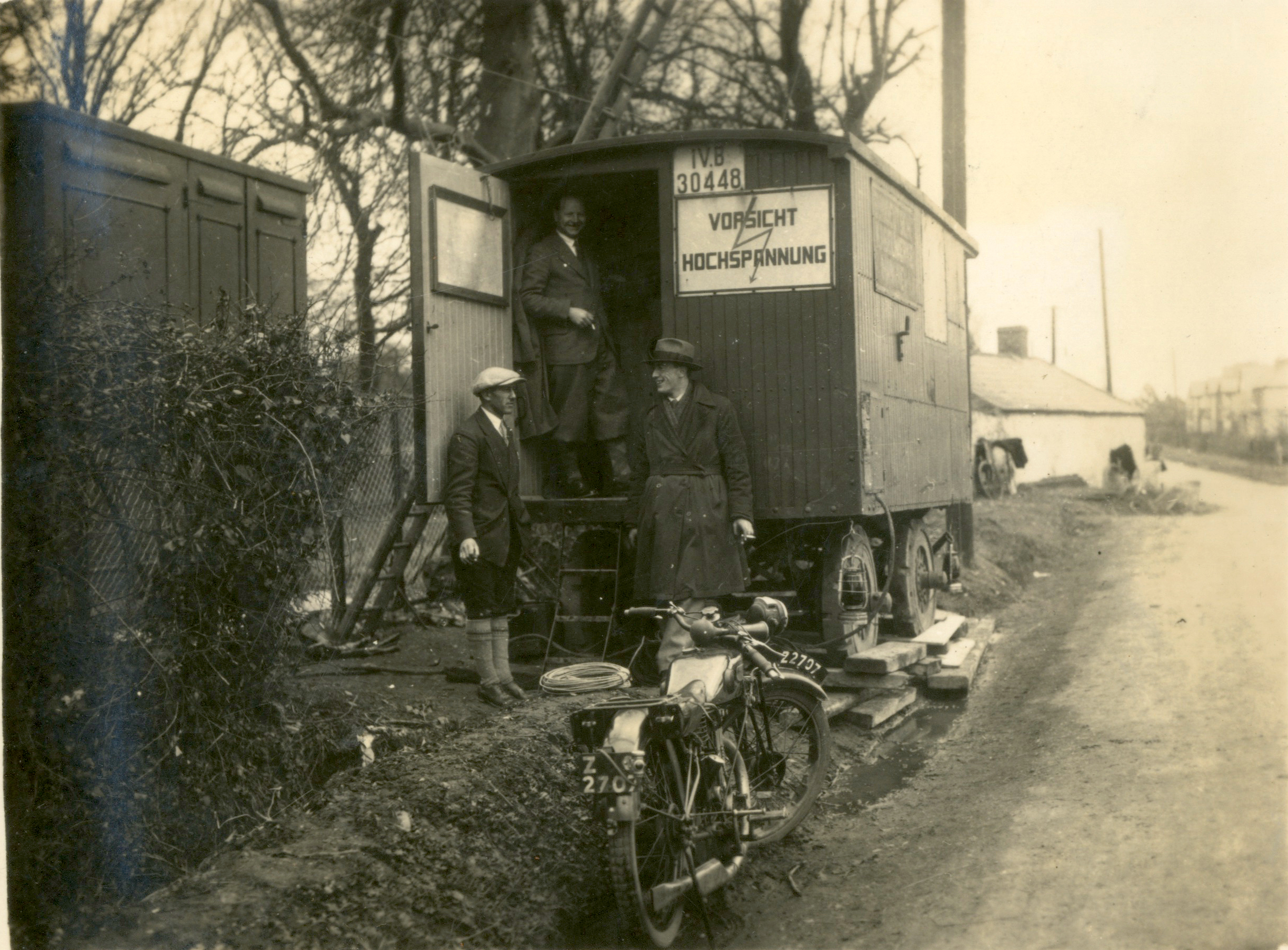 Black-and-white photograph of a temporary high-voltage electricity installation located alongside a rural roadway. Three utility workers are standing at the entrance to a mobile substation or control cabin mounted on a trailer.