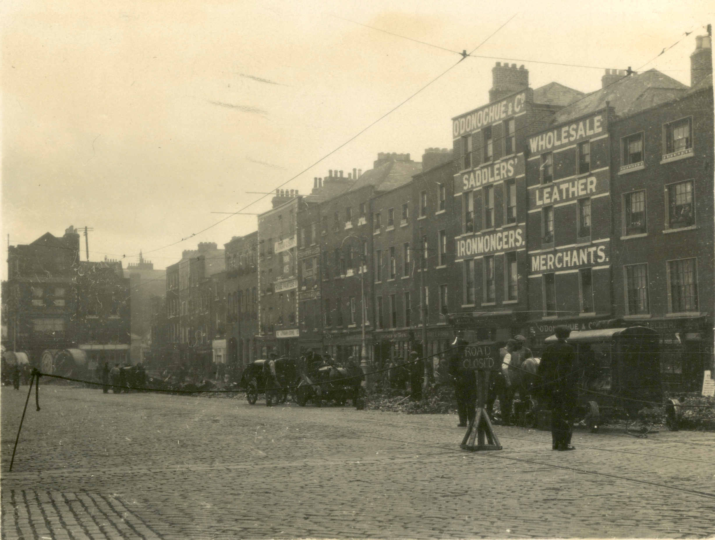 Black-and-white photograph of a city street during the installation of an electricity distribution network. A rope is stretched accross the roadway blocking public access while workers and vehicles are gathered beside excavation works along the street.