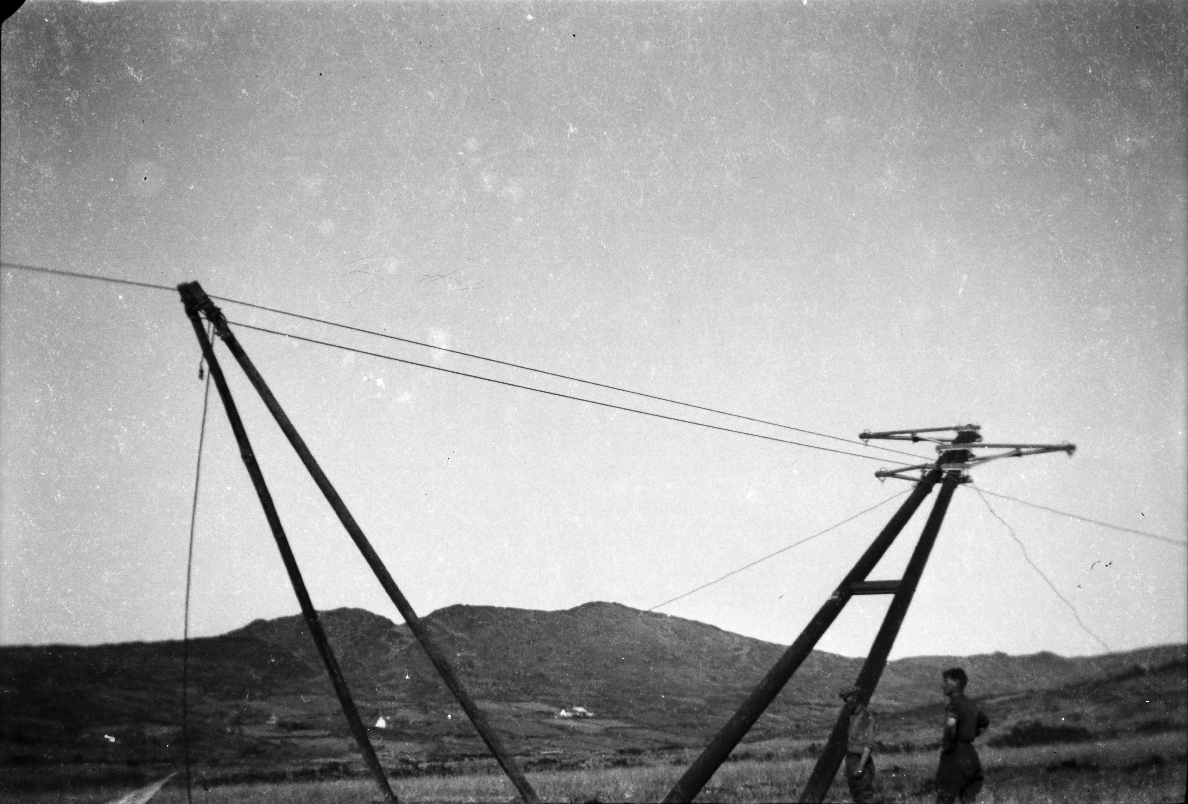 Black-and-white photograph showing twin wooden electricity support structures connected by overhead conductors, set against a mountainous rural landscape with two workers visible below.