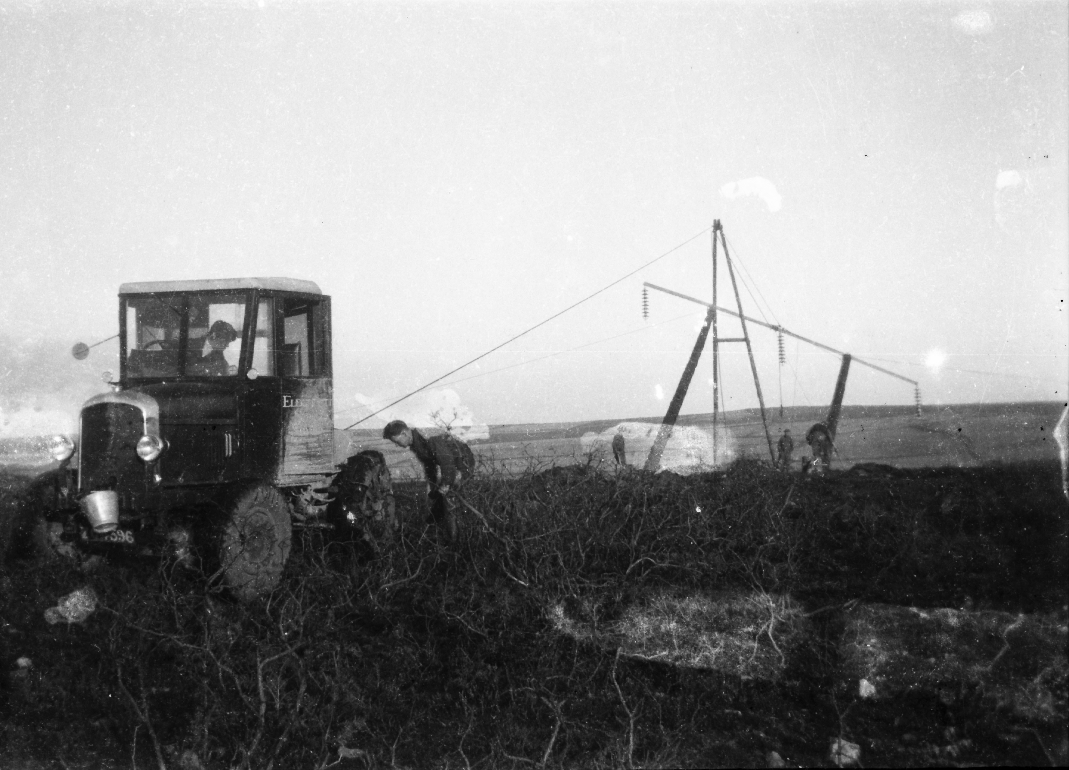 Black-and-white photograph showing an ESB tractor being used to erect an electricity line structure, with overhead conductors, support wires and several workers visible in the background.