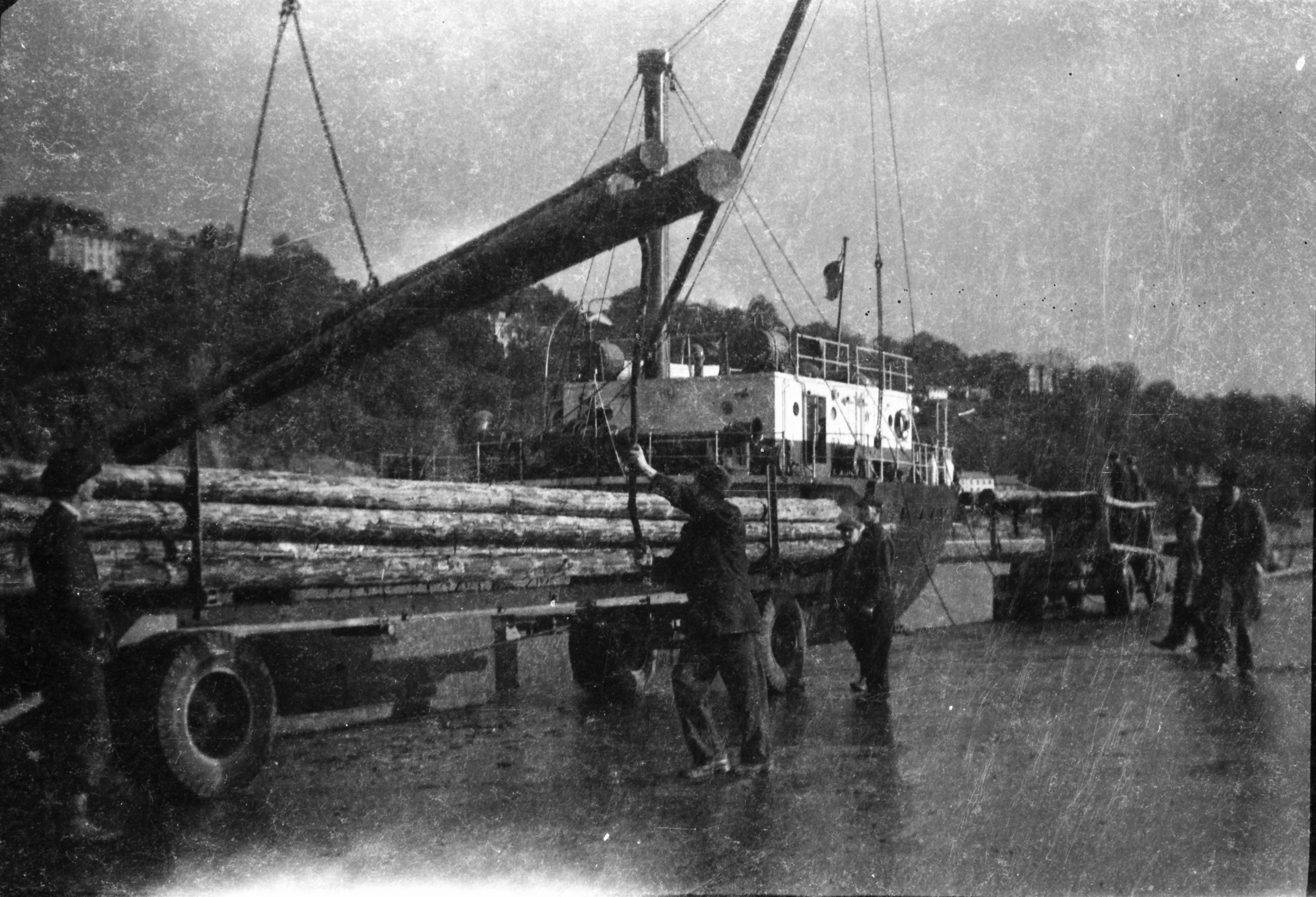 Black-and-white photograph showing dock workers transferring electricity poles from a ship to a lorry using a crane, with stacks of poles already secured on the vehicle.