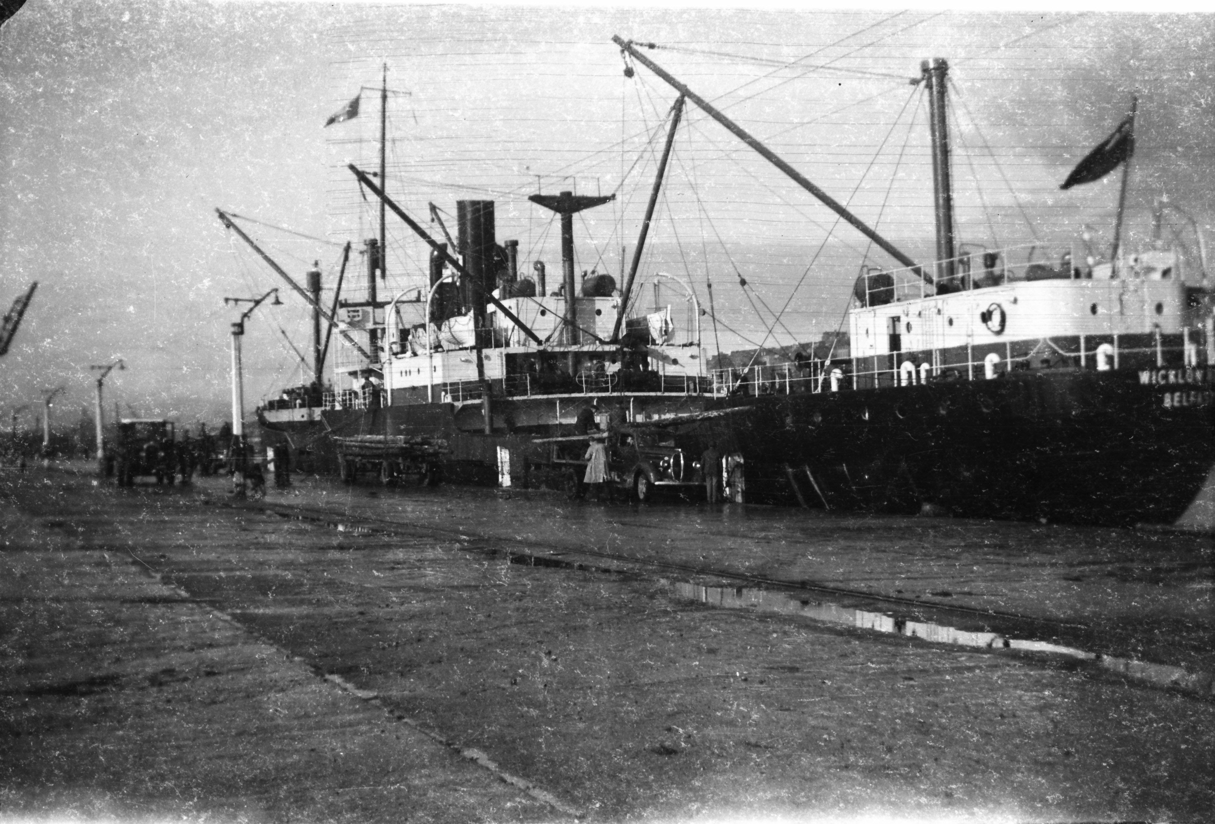 Black-and-white photograph showing a cargo ship berthed at a port, with cranes positioned for unloading electricity poles and several vehicles on the quayside.