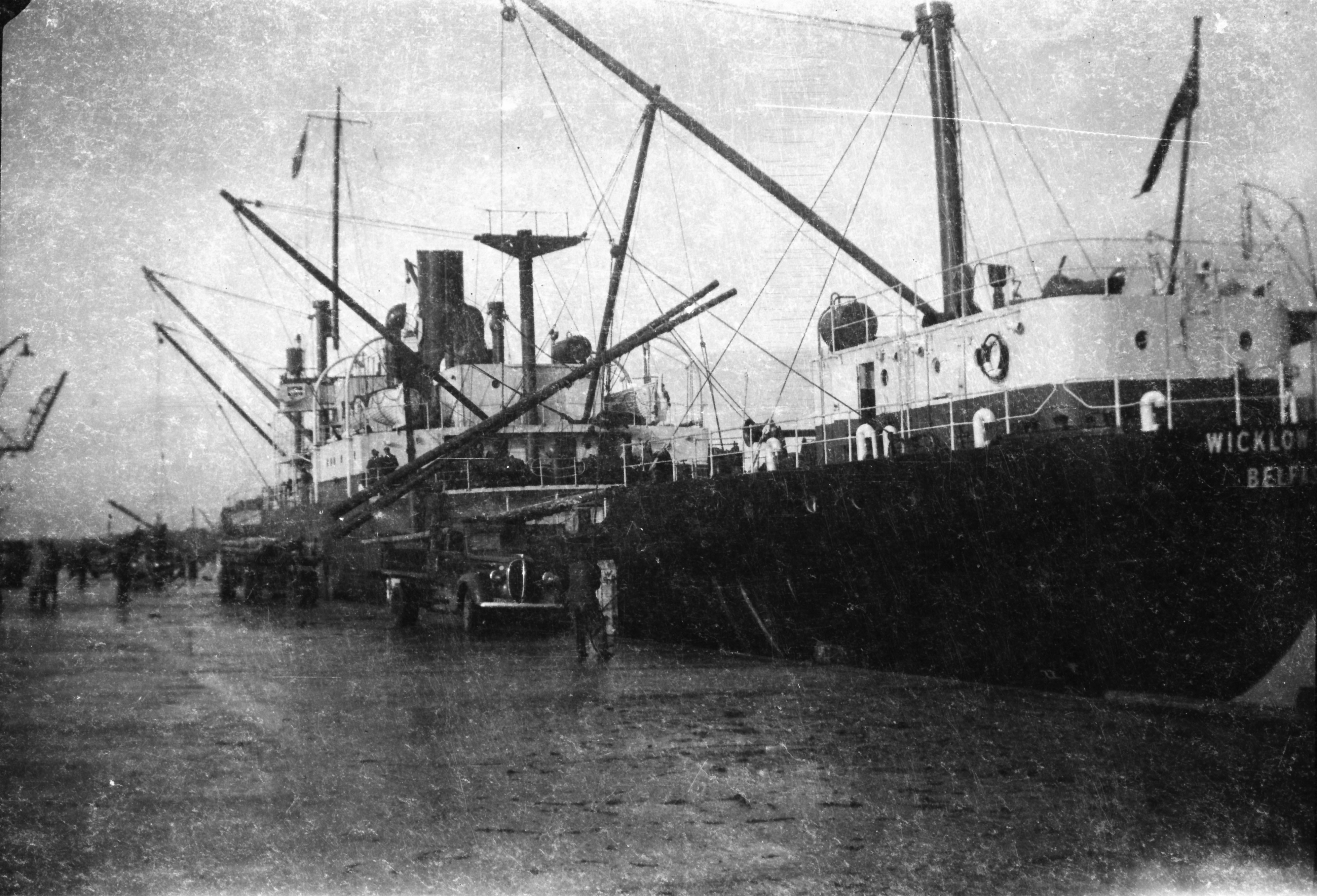 Black-and-white photograph showing electricity poles being unloaded from a cargo ship using onboard cranes, with workers and vehicles positioned on the quayside below.