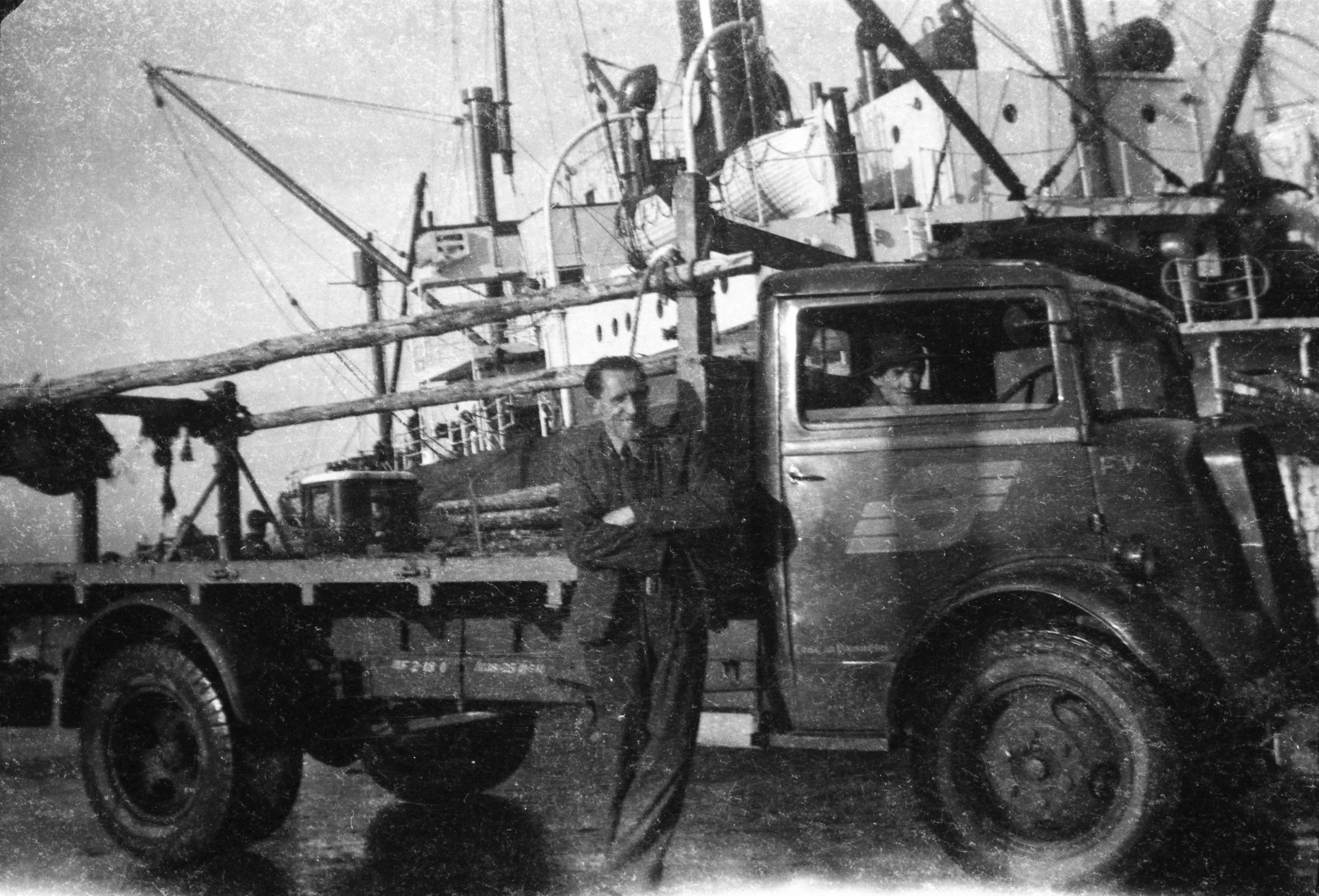 Black-and-white photograph showing a flatbed lorry and two workers on a dockside, with a cargo vessel and unloading equipment in the background.