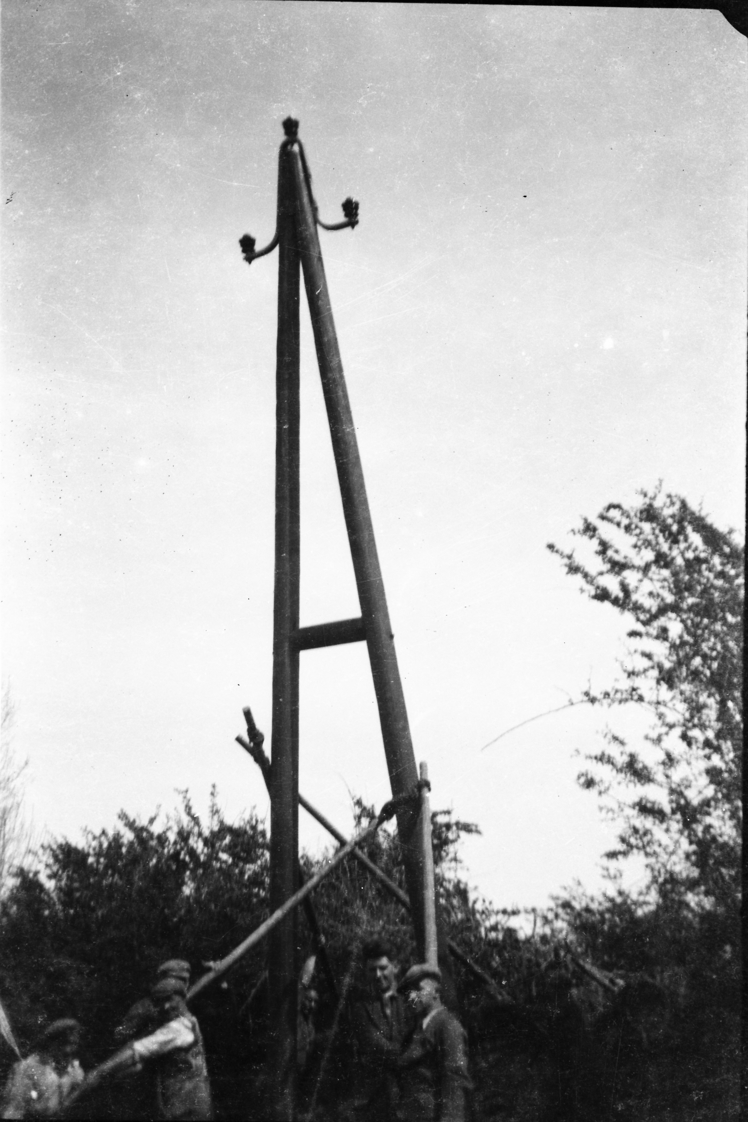 Black-and-white photograph showing several workers erecting a wooden H-frame electricity support structure, using guide poles to steady the framework during installation.