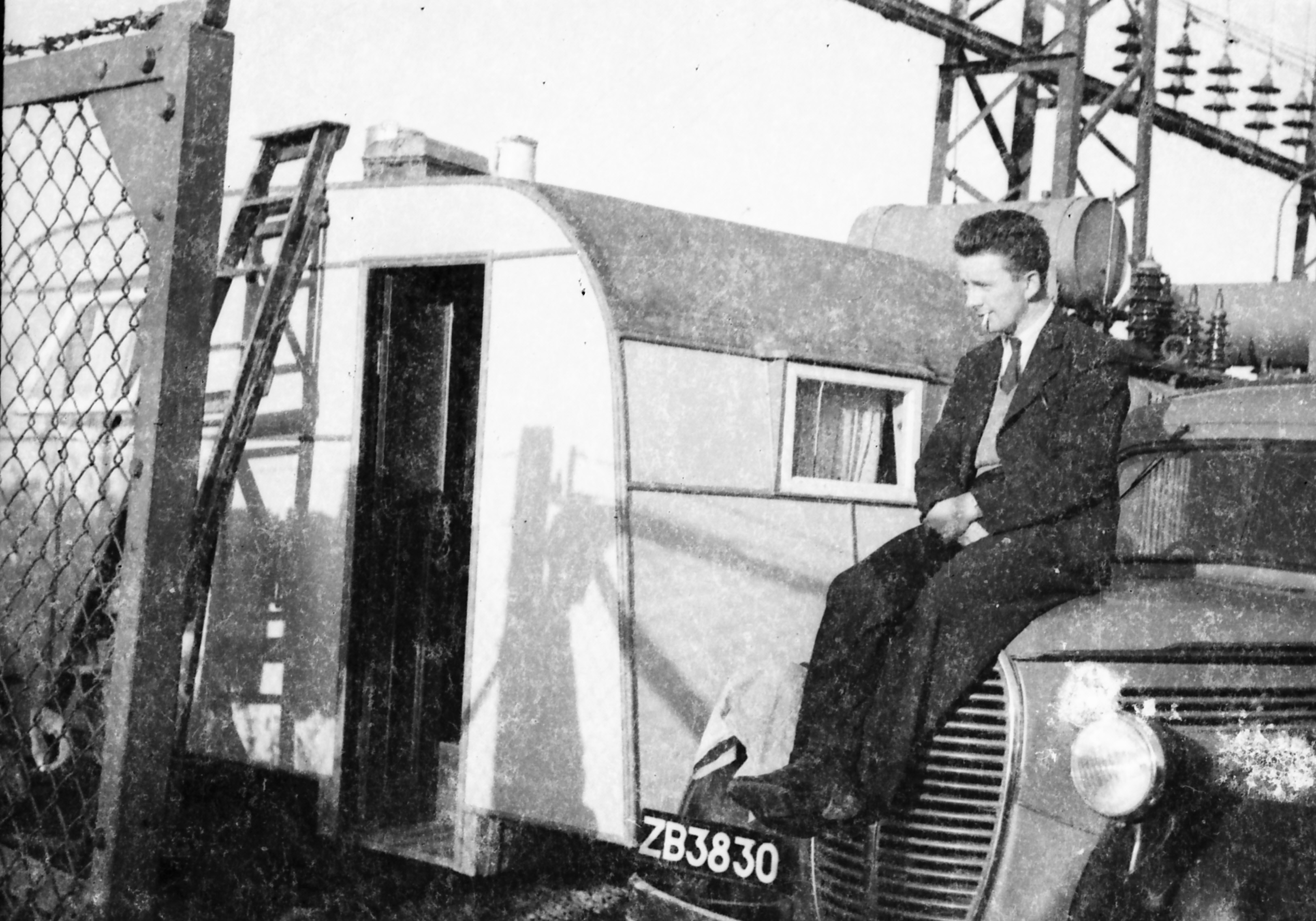 Black-and-white photograph an ESB worker seated on the front of a vehicle parked next to a caravan at an electricity installation site, with transmission structures visible behind him.