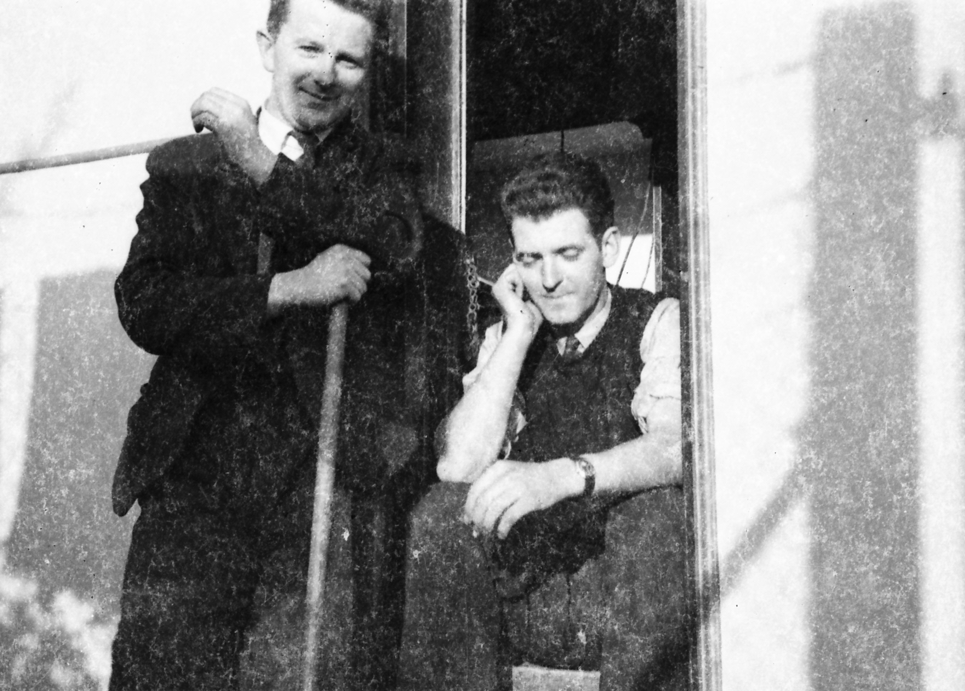 Black-and-white photograph showing Jerry Corbett seated in a doorway while a colleague stands beside him holding a long-handled tool.