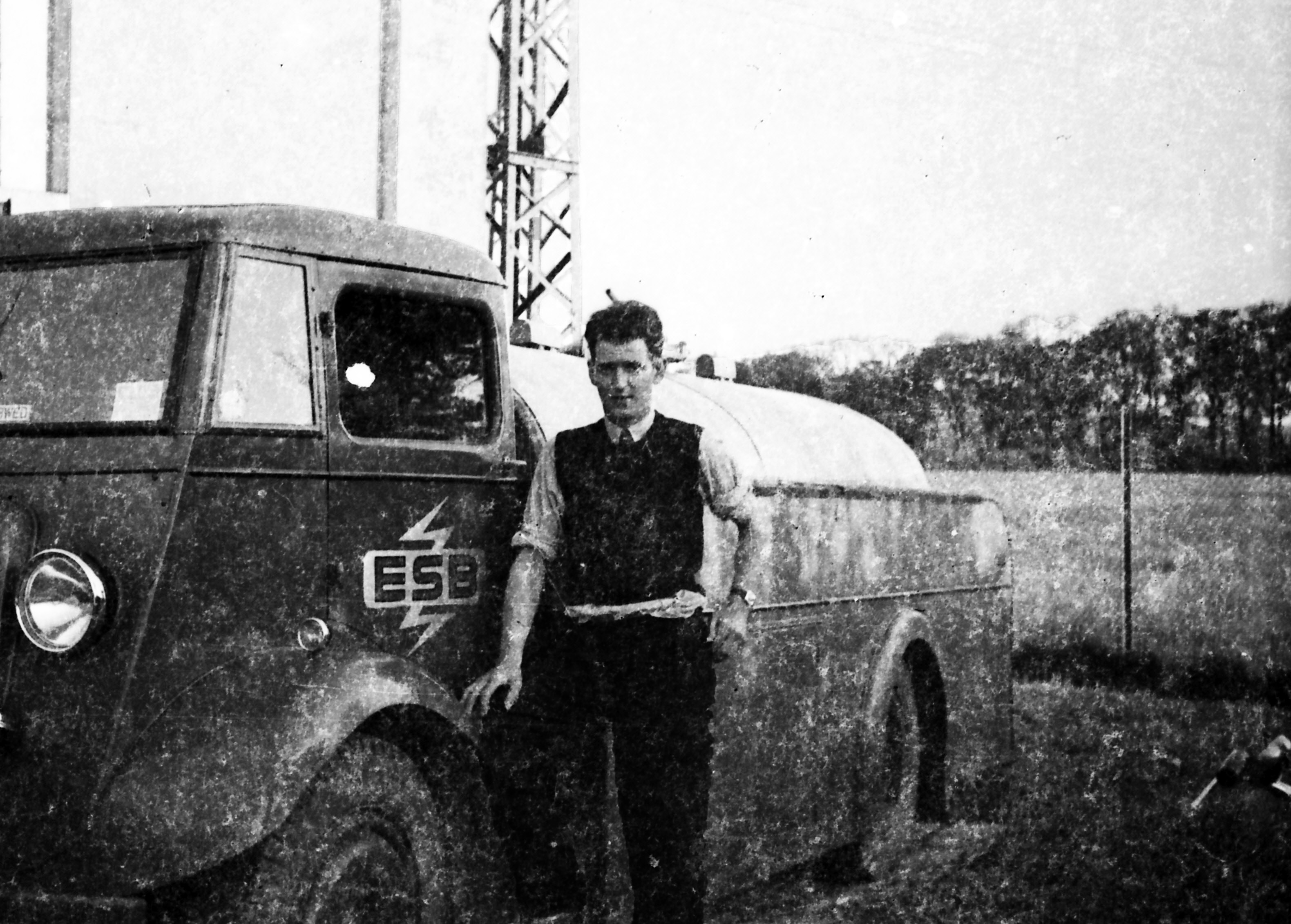 Black-and-white photograph showing Jerry Corbett standing next to an ESB utility vehicle bearing the ESB logo, with electricity infrastructure and open countryside behind him.