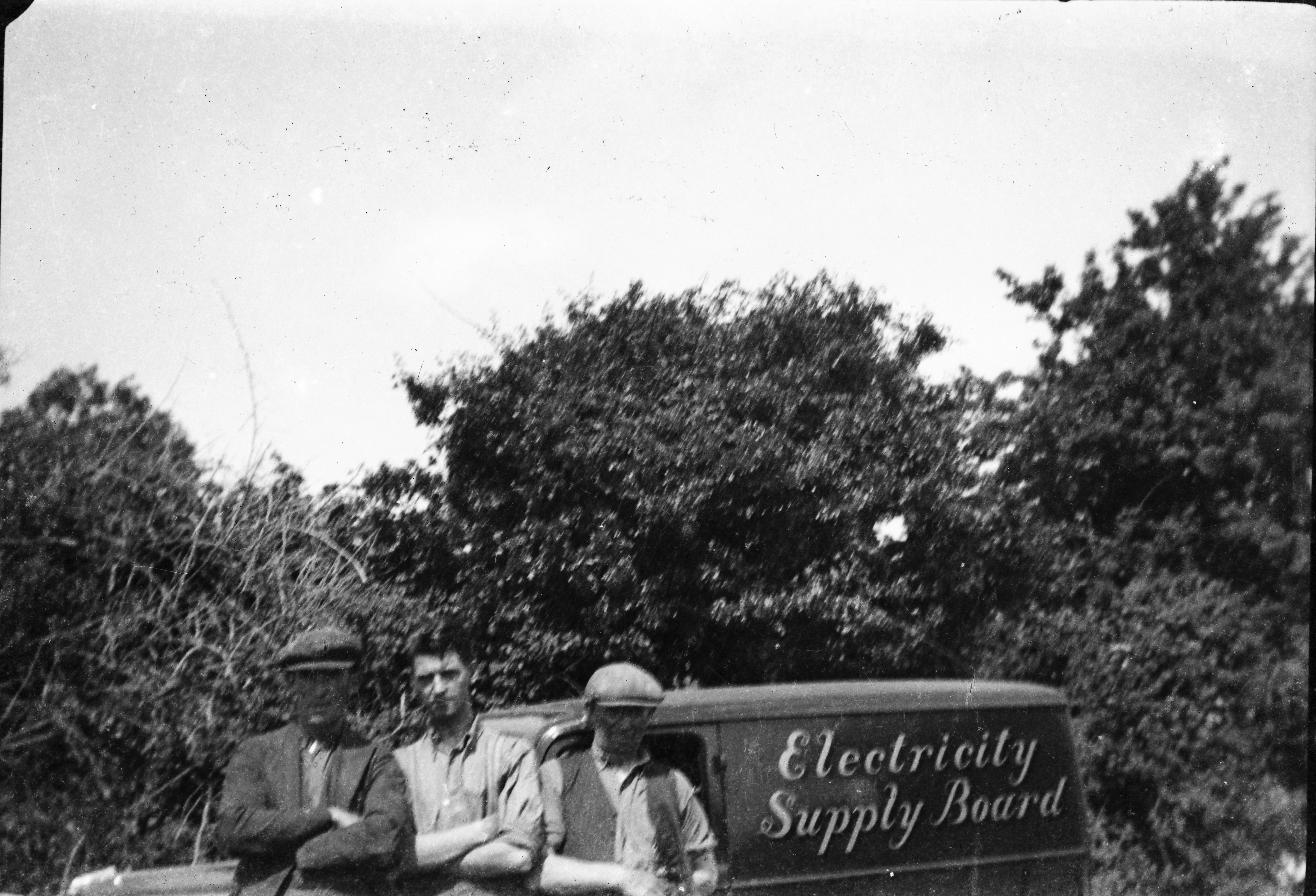 Black-and-white photograph showing Jerry Corbett and two colleagues posing beside an Electricity Supply Board vehicle, with trees and hedgerows in the background.