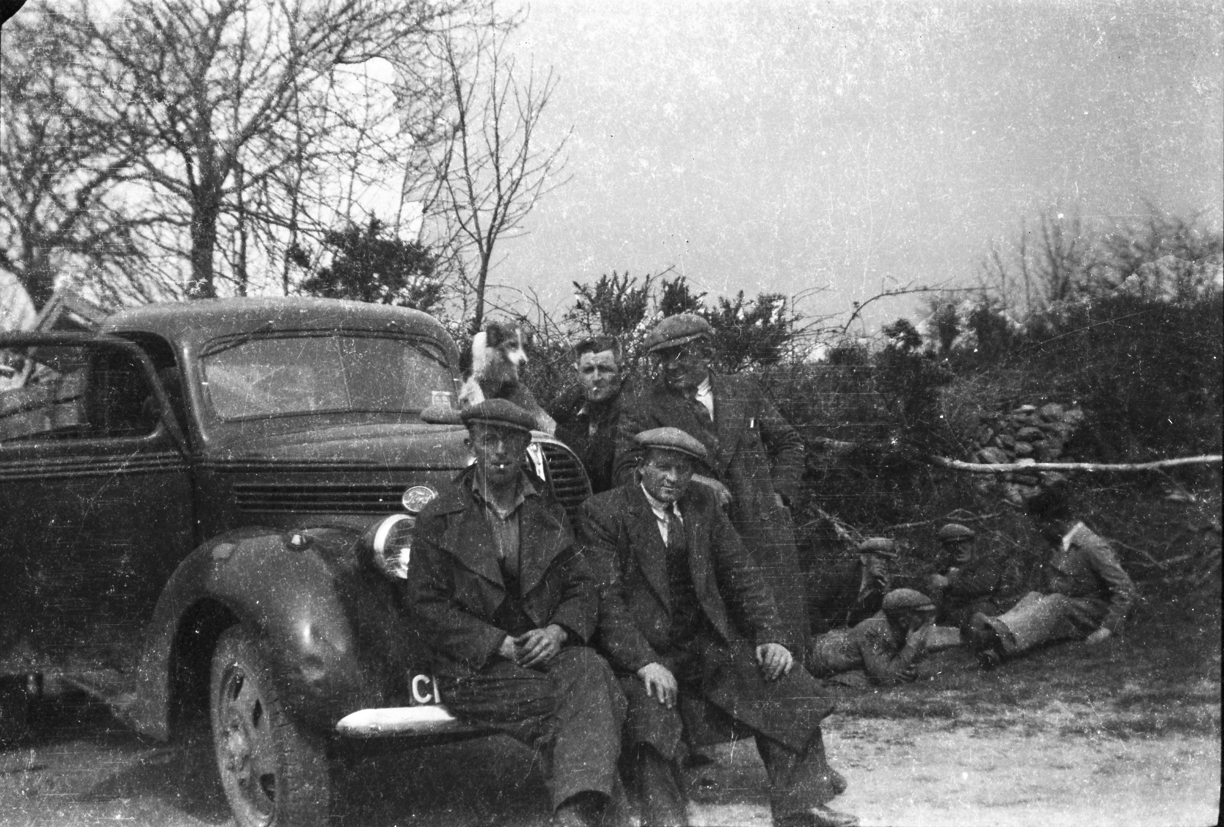 Black-and-white photograph showing workers taking a break beside a utility vehicle, with some posing for the camera and others seated on the ground near a stone wall.
