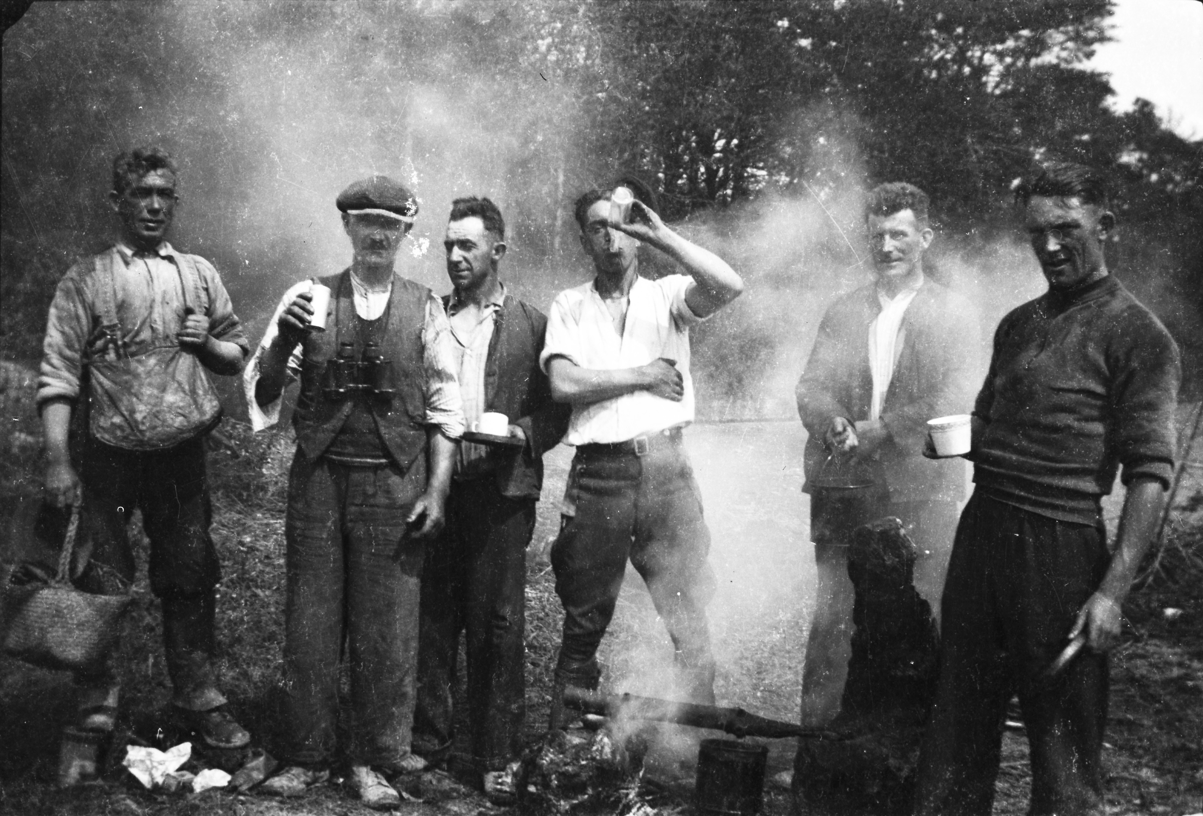 Black-and-white photograph showing six workers taking a break beside an open campfire, with cooking pots visible in the foreground and smoke drifting through the scene.