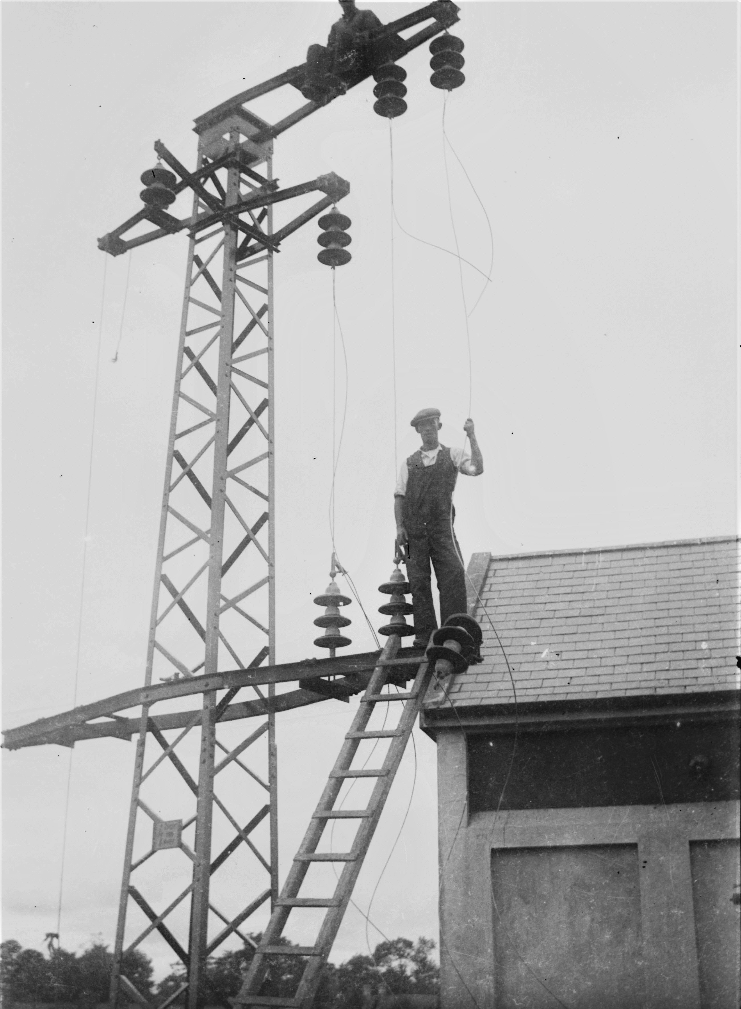 Black-and-white photograph showing workers installing insulators on a steel electricity support structure, with one worker standing on a ladder and another working near the top of the tower beside a building.
