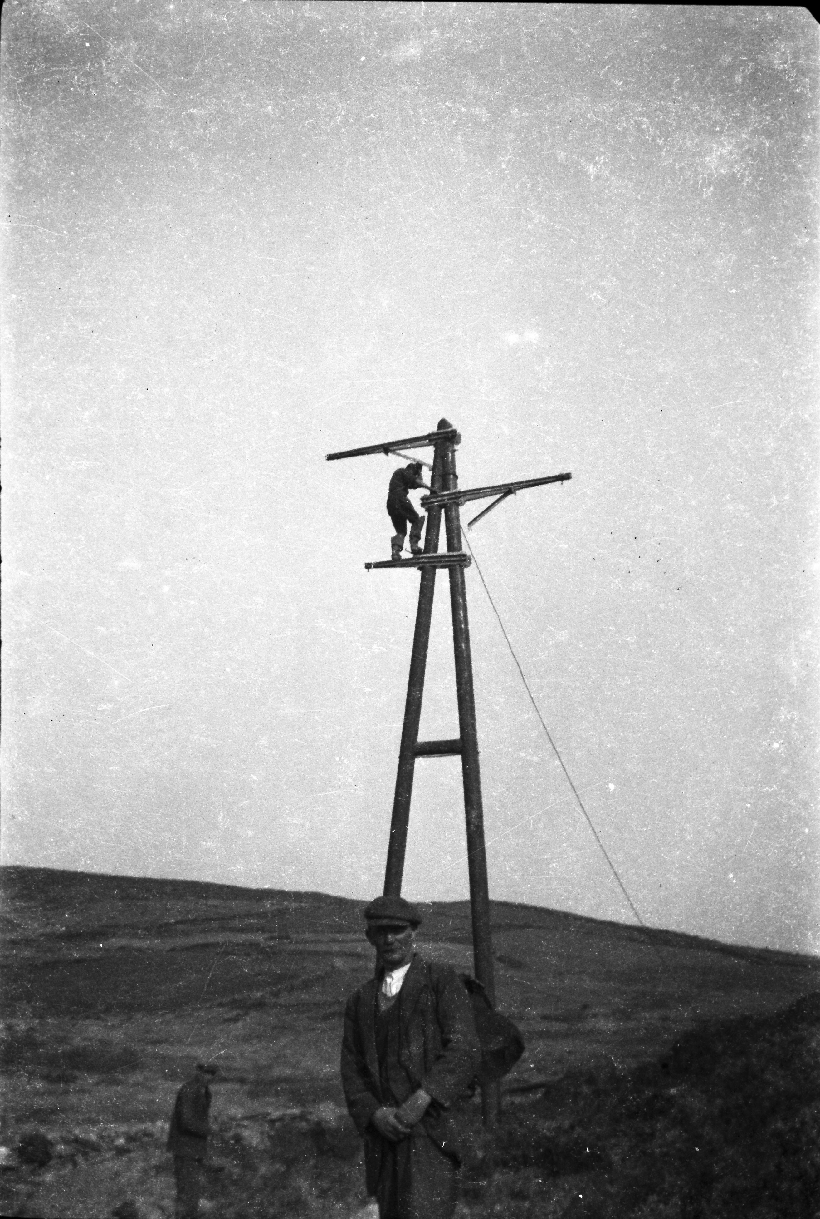 Black-and-white photograph showing a worker carrying out construction work high on a wooden electricity support structure, with a colleague standing at its base and hills visible in the background.