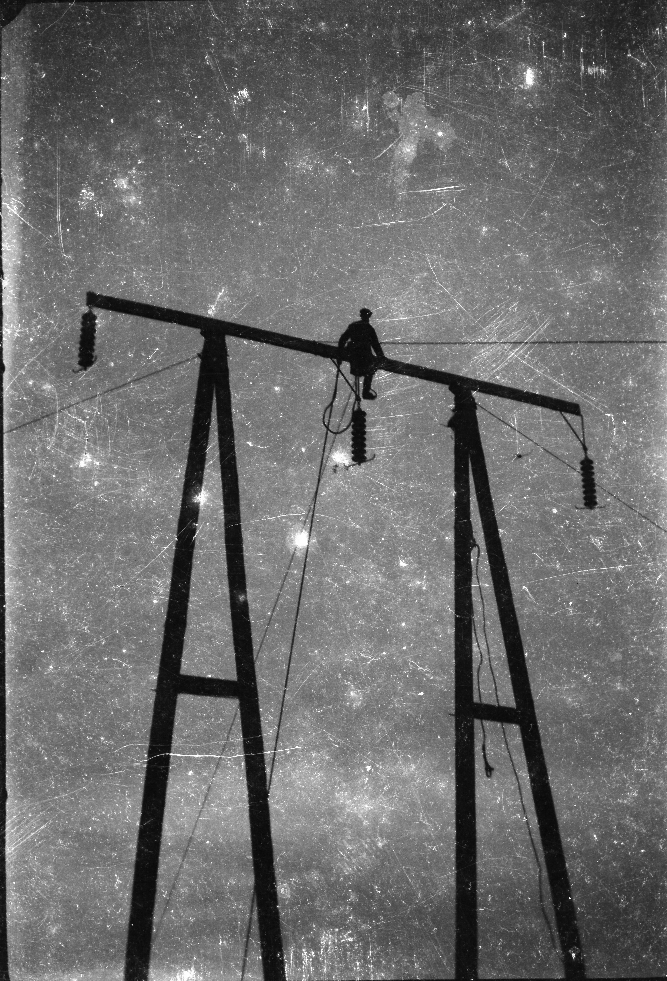 Black-and-white photograph showing a worker carrying out installation or maintenance work at the top of a wooden electricity support structure, silhouetted against the sky.