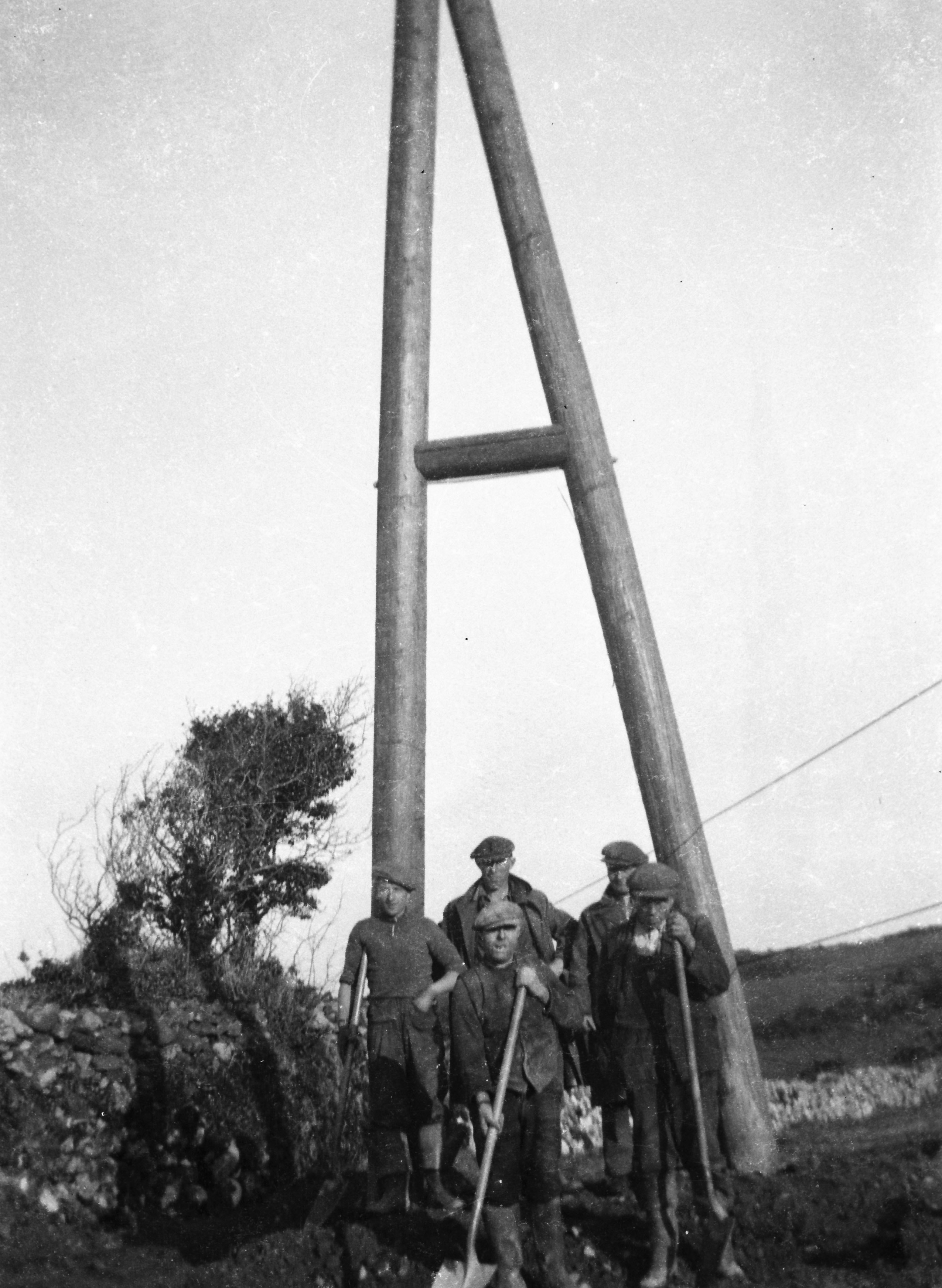 Black-and-white photograph showing workers gathered at the base of a wooden framed electricity support structure during power line construction, with stone walls and open countryside visible nearby.