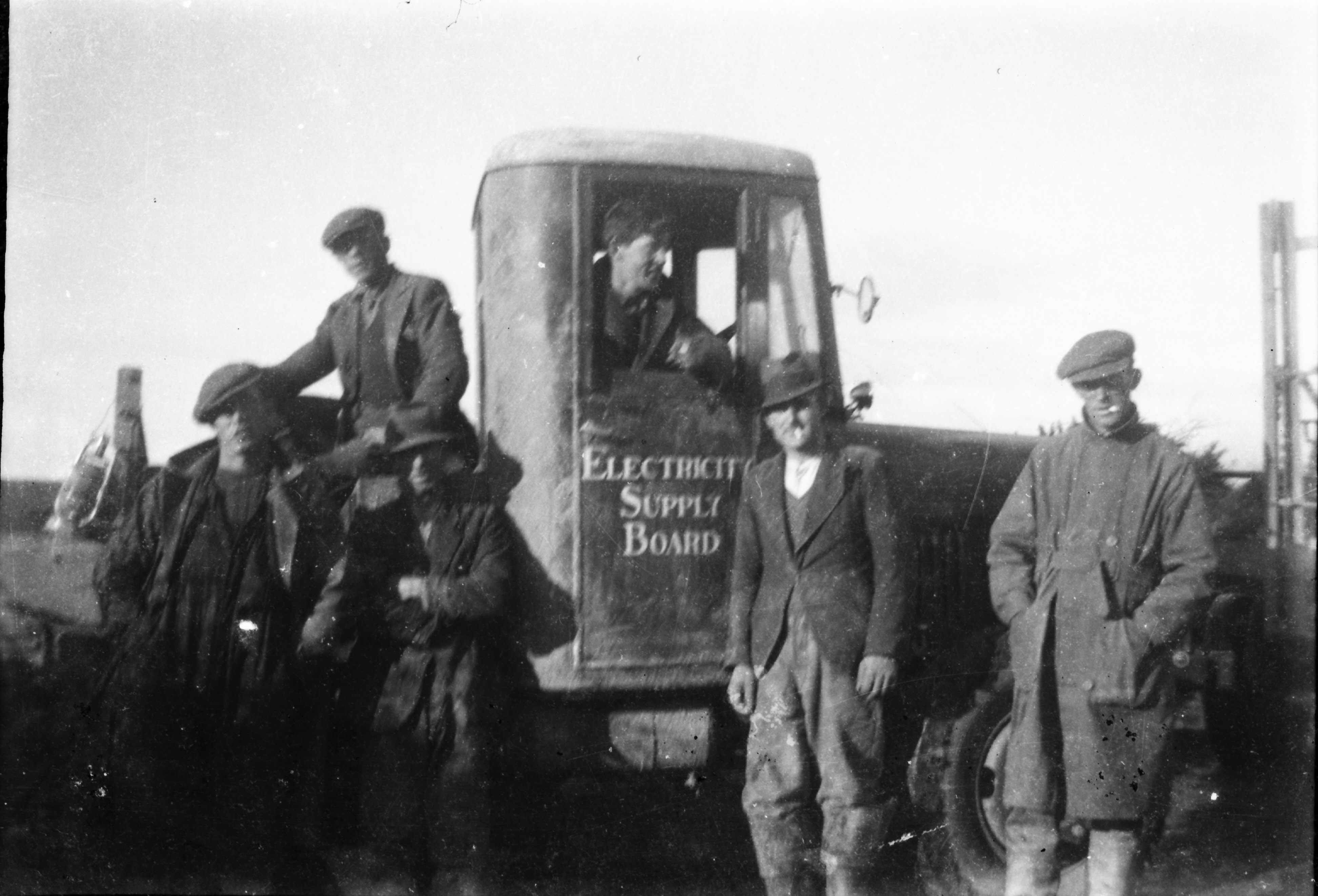 Black-and-white photograph showing workers gathered around an Electricity Supply Board vehicle at a rural work site, with one person visible through the cab window and others standing alongside.