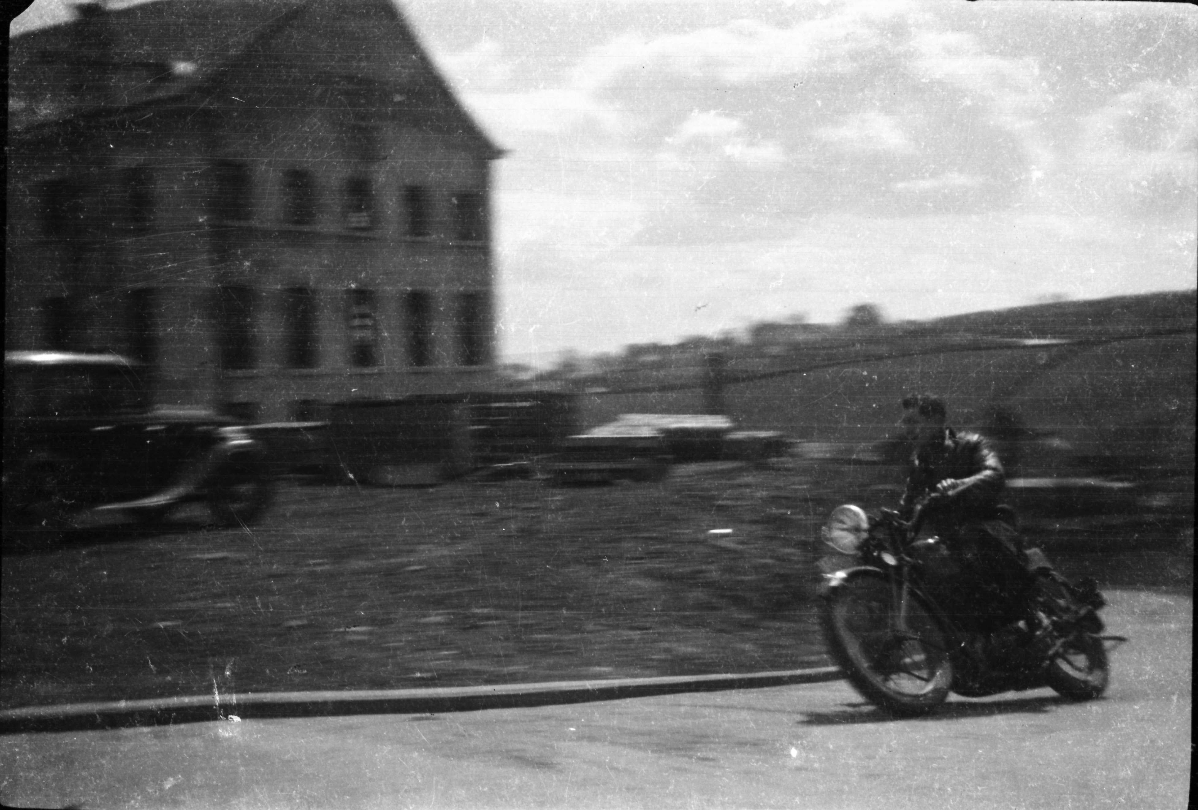 Black-and-white photograph capturing a motorcyclist travelling past a large building and adjacent yard, with cars parked nearby and motion blur visible across the scene.