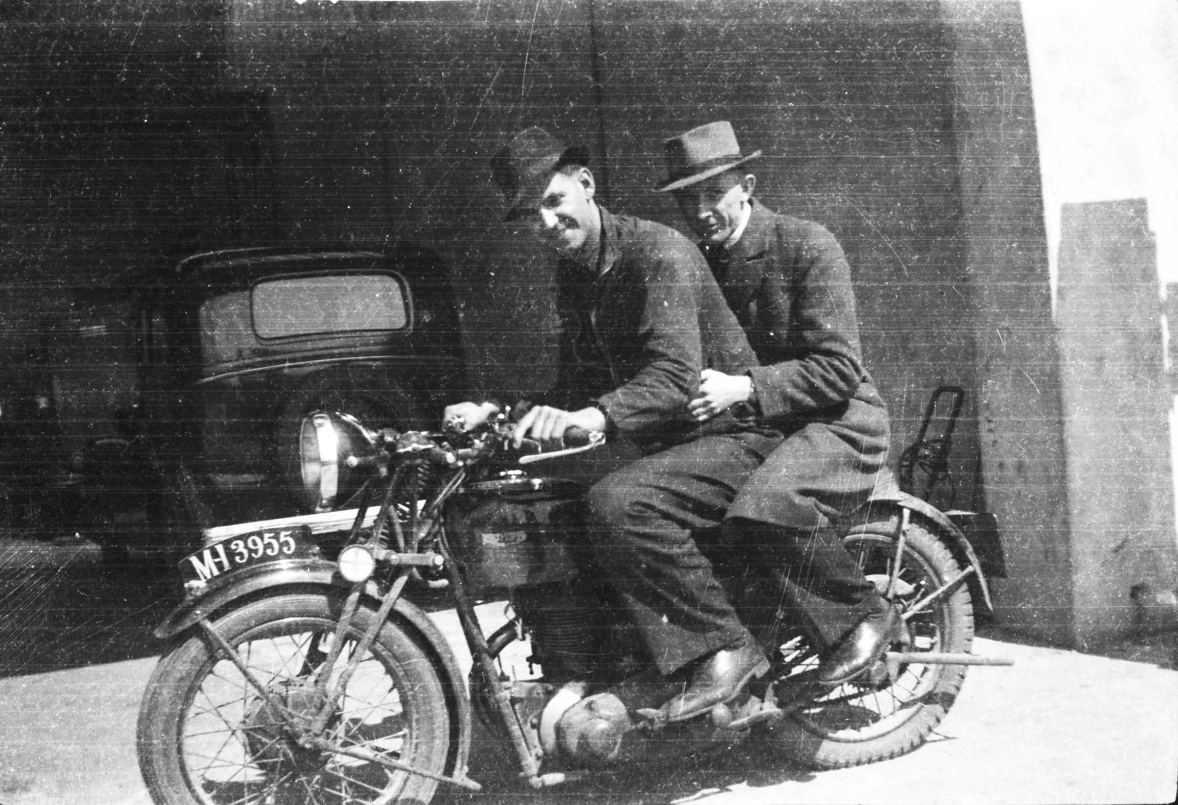 Black-and-white photograph showing Jerry Corbett sitting on a motorcycle with another person seated behind him, in front of a large entryway with a parked car nearby.
