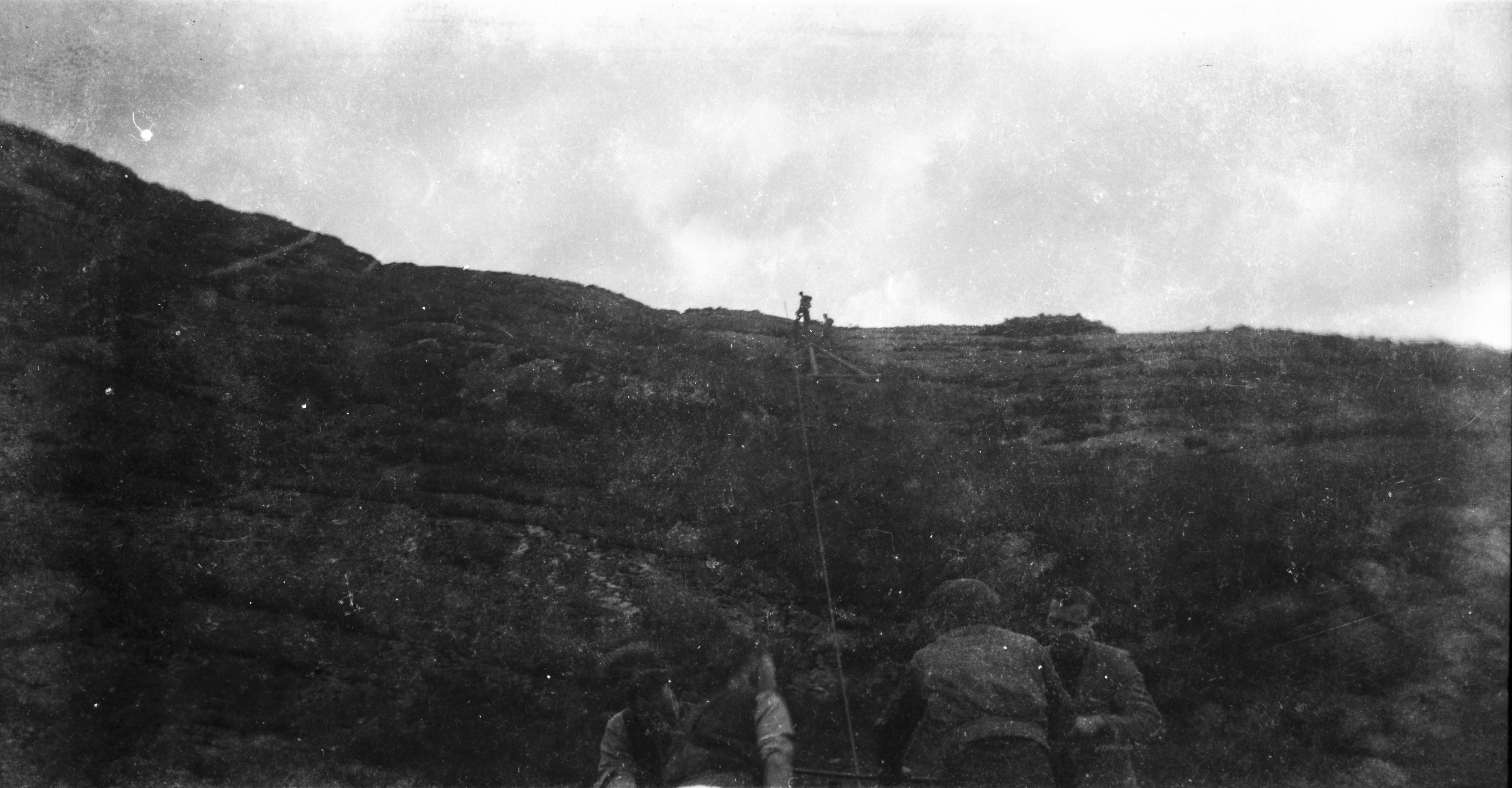 Black-and-white photograph showing workers on a rugged hillside pulling a rope line from below while colleagues stand near the ridge above.