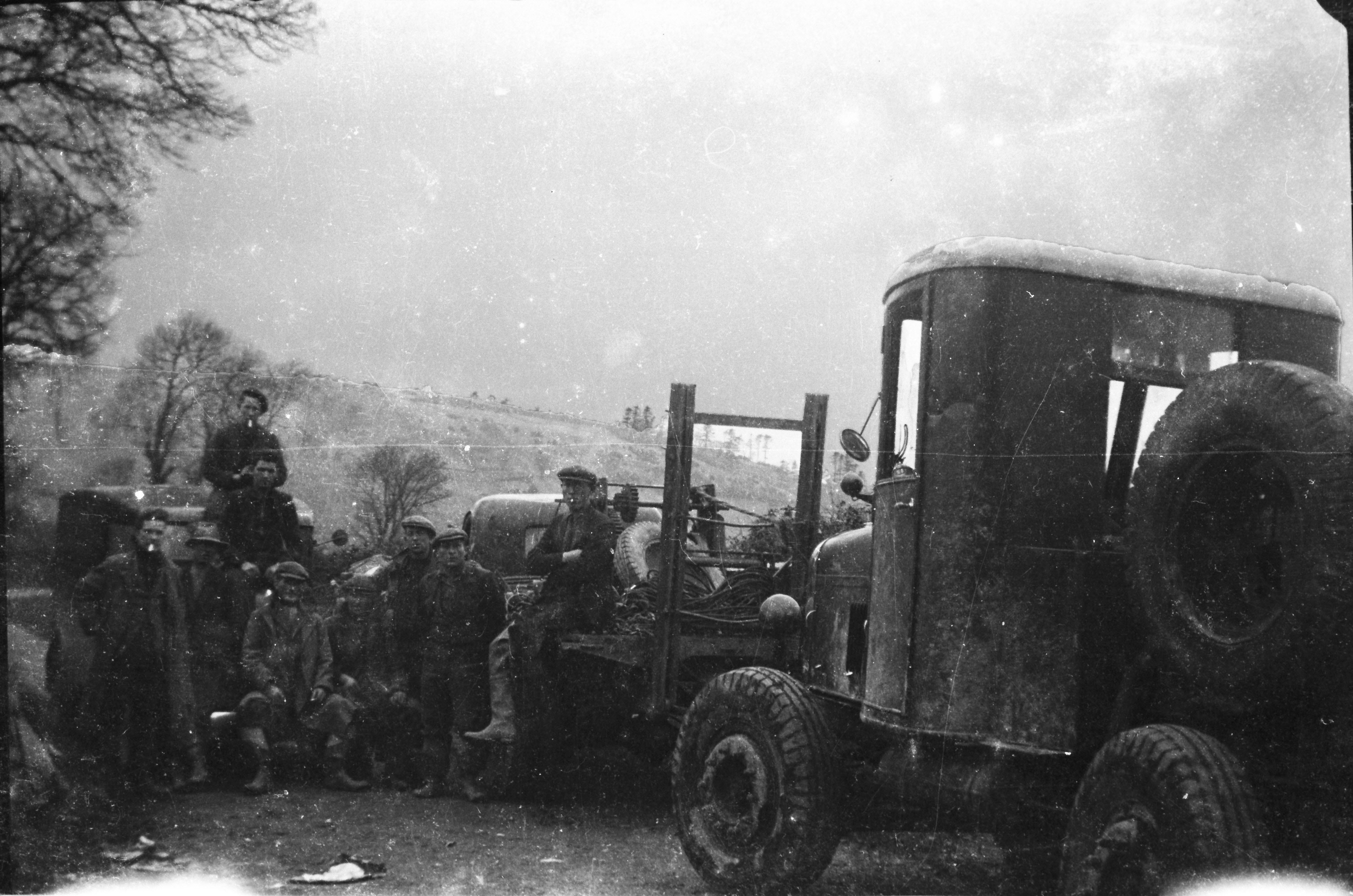 Black-and-white photograph showing workers posing beside utility vehicles at a rural work site, with tools, cable reels and equipment visible on one vehicle.
