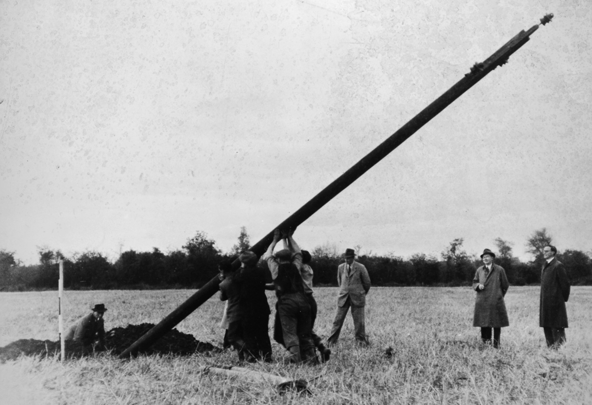 Black-and-white photograph showing workers raising a wooden electricity pole into position in an open field while several observers stand nearby.