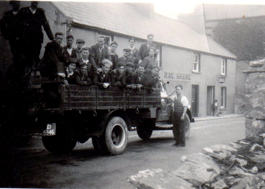 Group of ESB workers standing and sitting on the back of a lorry parked on a village street, with buildings visible in the background.