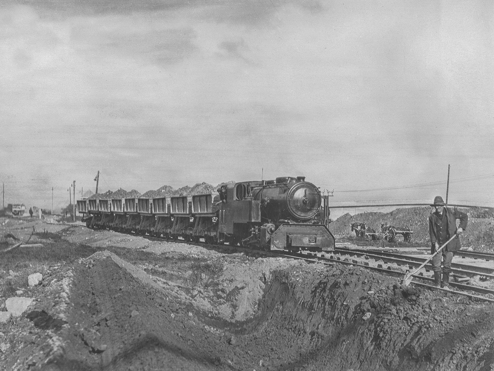 Black-and-white photograph showing a construction railway train transporting earth along a temporary track, with excavation works and workers visible in the foreground.