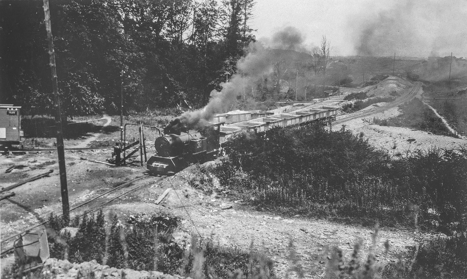 Black-and-white photograph showing a steam construction train hauling loaded wagons along a temporary railway through a large earthworks site, with smoke rising from the locomotive.