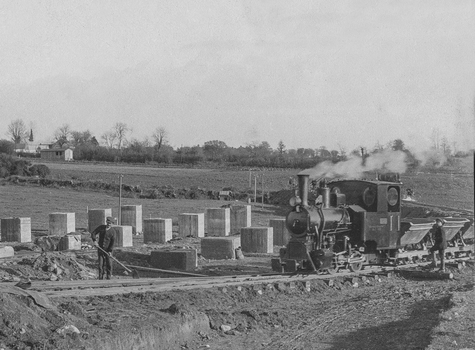 Black-and-white photograph showing a steam construction train hauling tipper wagons through an excavation site, with concrete foundation blocks and earthworks visible alongside the track.