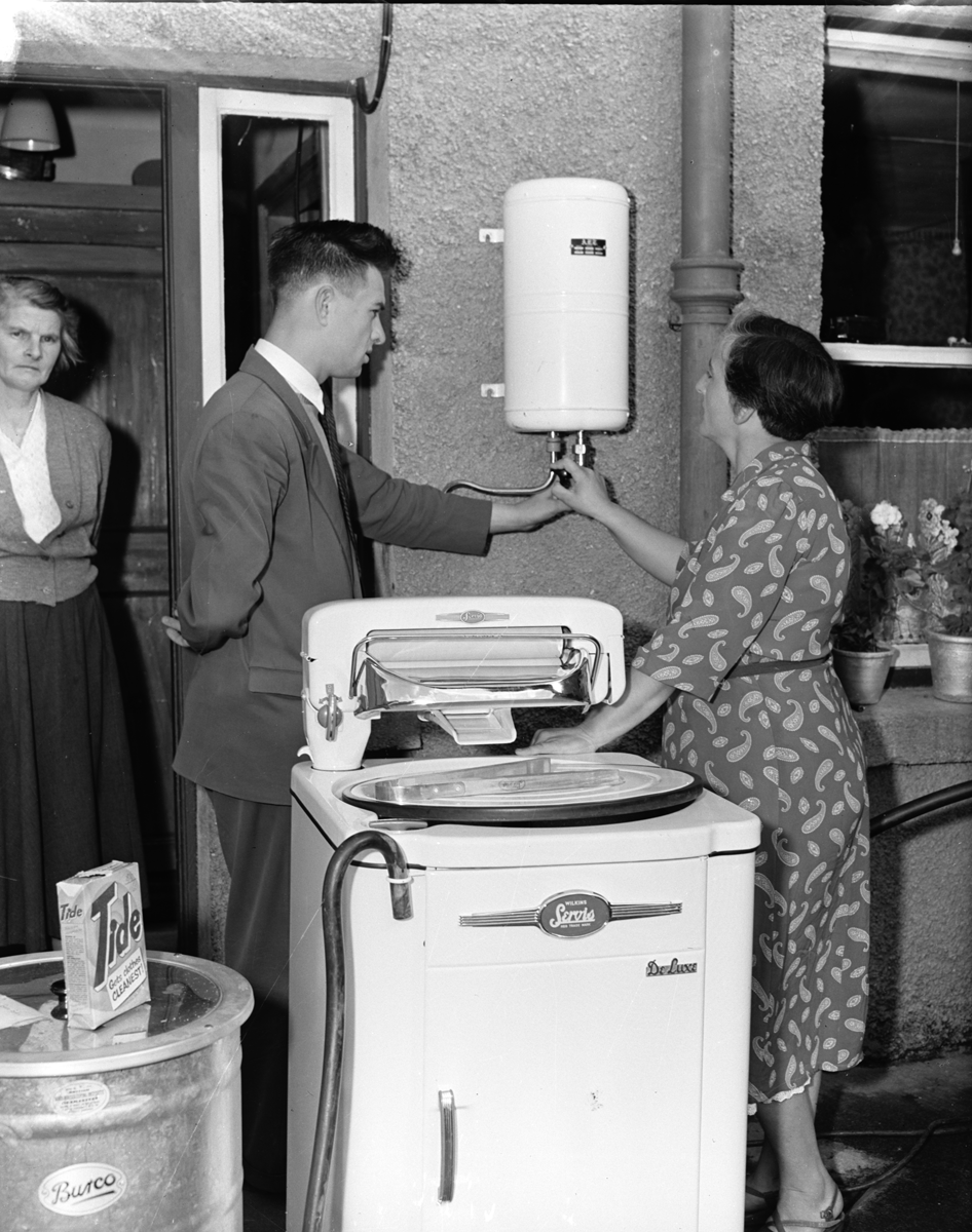 Black-and-white photograph showing a demonstration of an electric washing machine and water heater at an outdoor rural electrification exhibition, with attendees being shown how the appliances operate.