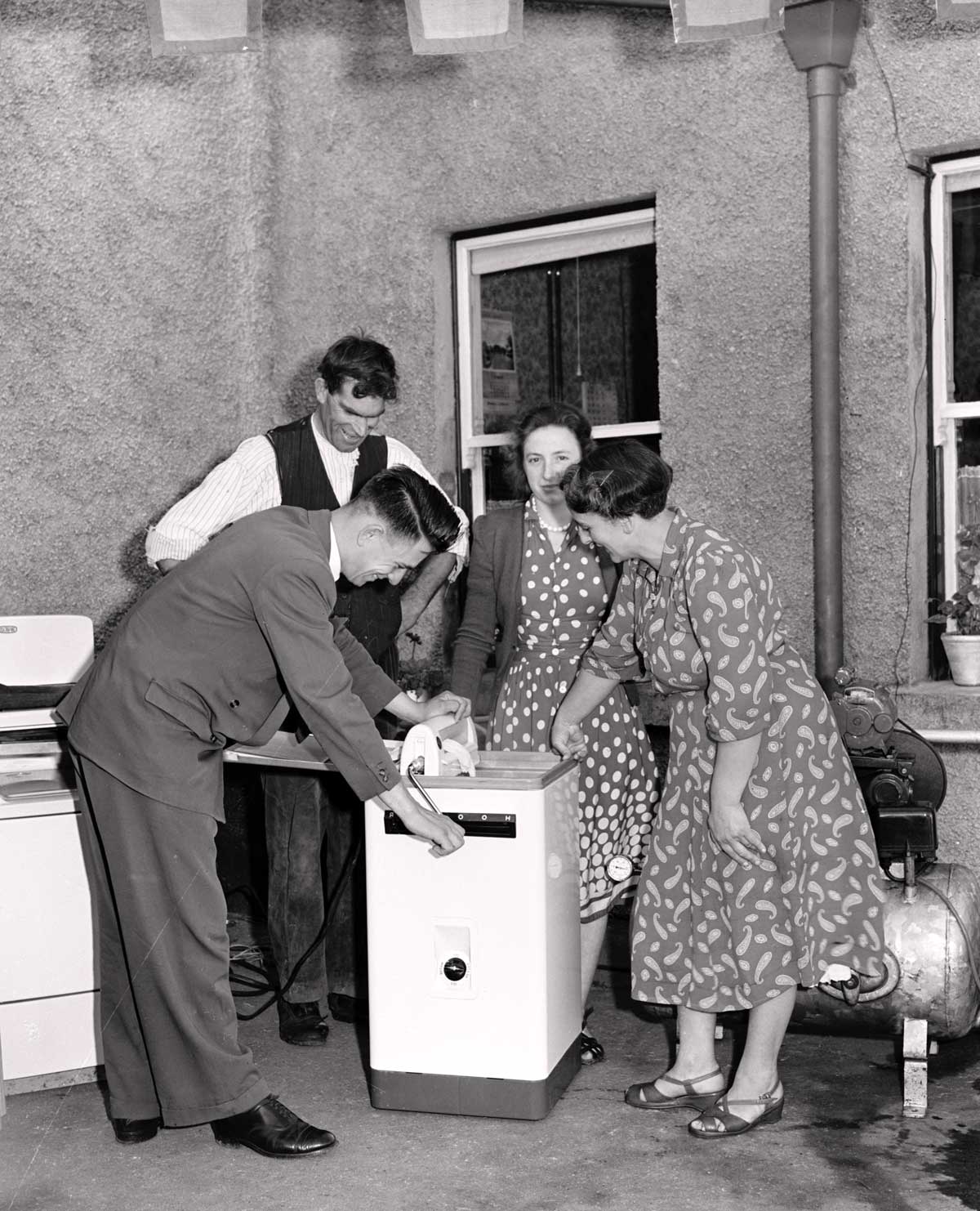 Black-and-white photograph showing a demonstration of an electric washing machine at an outdoor rural electrification exhibition, with attendees gathered around the appliance outside a building.