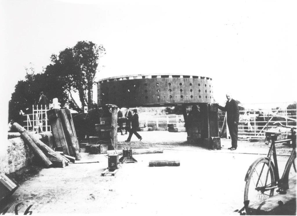 Large circular generator component raised on transport supports beside a rural road, with several people standing nearby and a bicycle visible in the foreground.