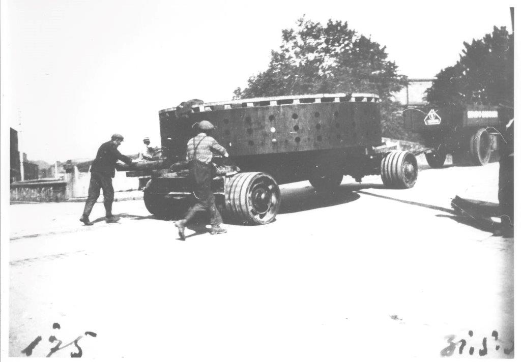 Black-and-white photograph showing a large circular generator component on a heavy transport platform with multiple wheels, as workers guide it through a road junction during its journey.