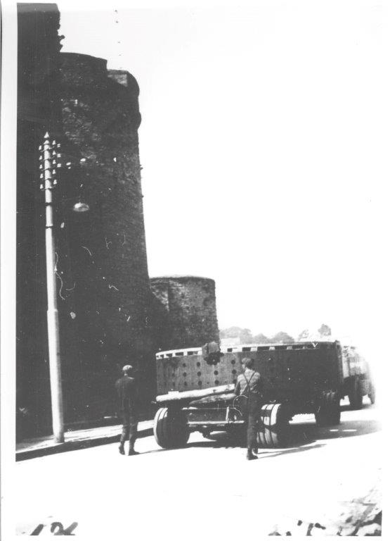 Black and white photograph of a large cylindrical generator component on a transport vehicle travelling along a city street beside a stone castle wall, with two people walking alongside the load.