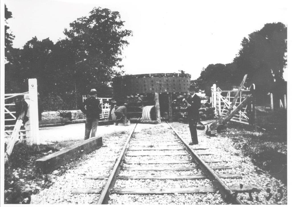 Black-and-white photograph showing a heavy generator component being transported across a railway crossing, with workers and onlookers positioned around the load and gates on either side of the track.