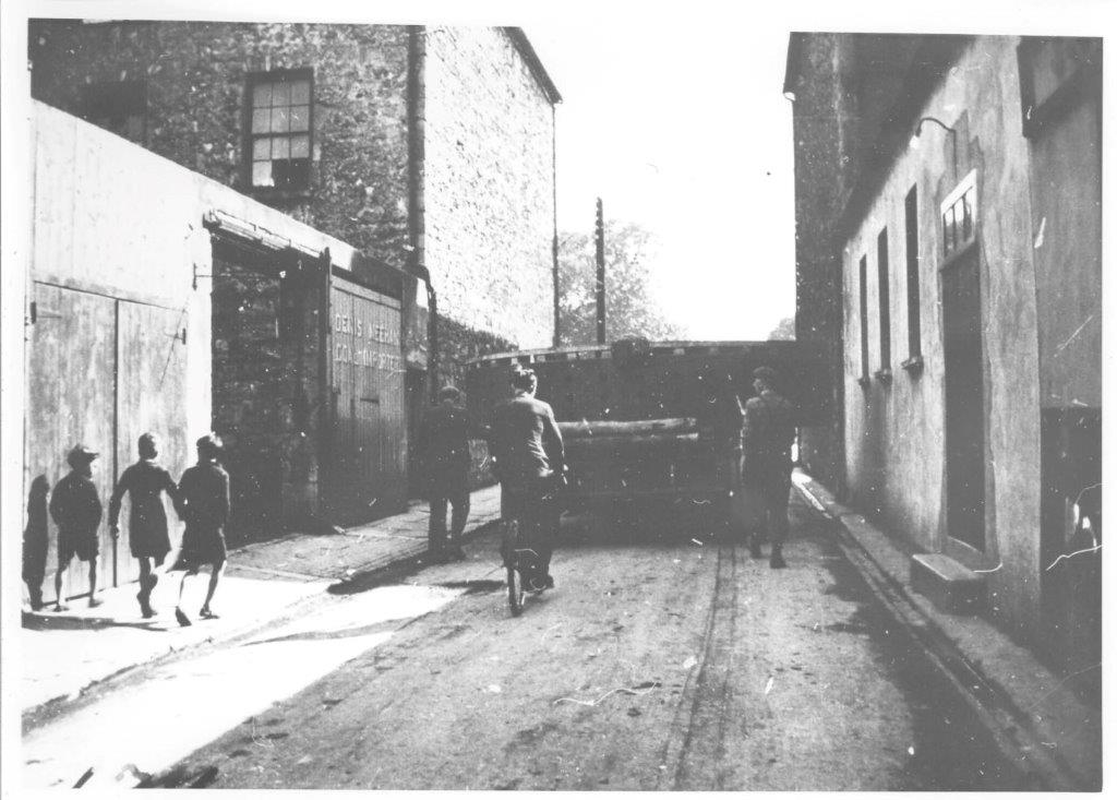 Black-and-white photograph showing a heavy generator component being transported along a confined urban street, passing between rows of buildings with pedestrians and a cyclist nearby.
