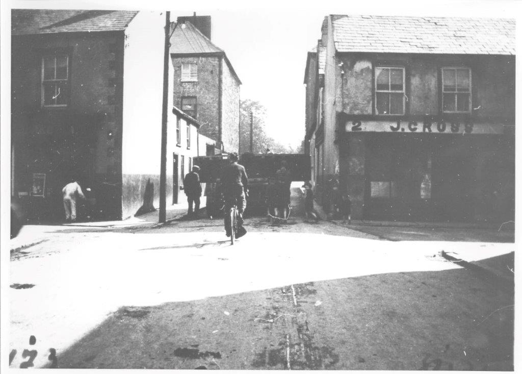 Black-and-white photograph showing a heavy generator component being transported along a confined urban street, passing between rows of buildings with pedestrians and a cyclist nearby.
